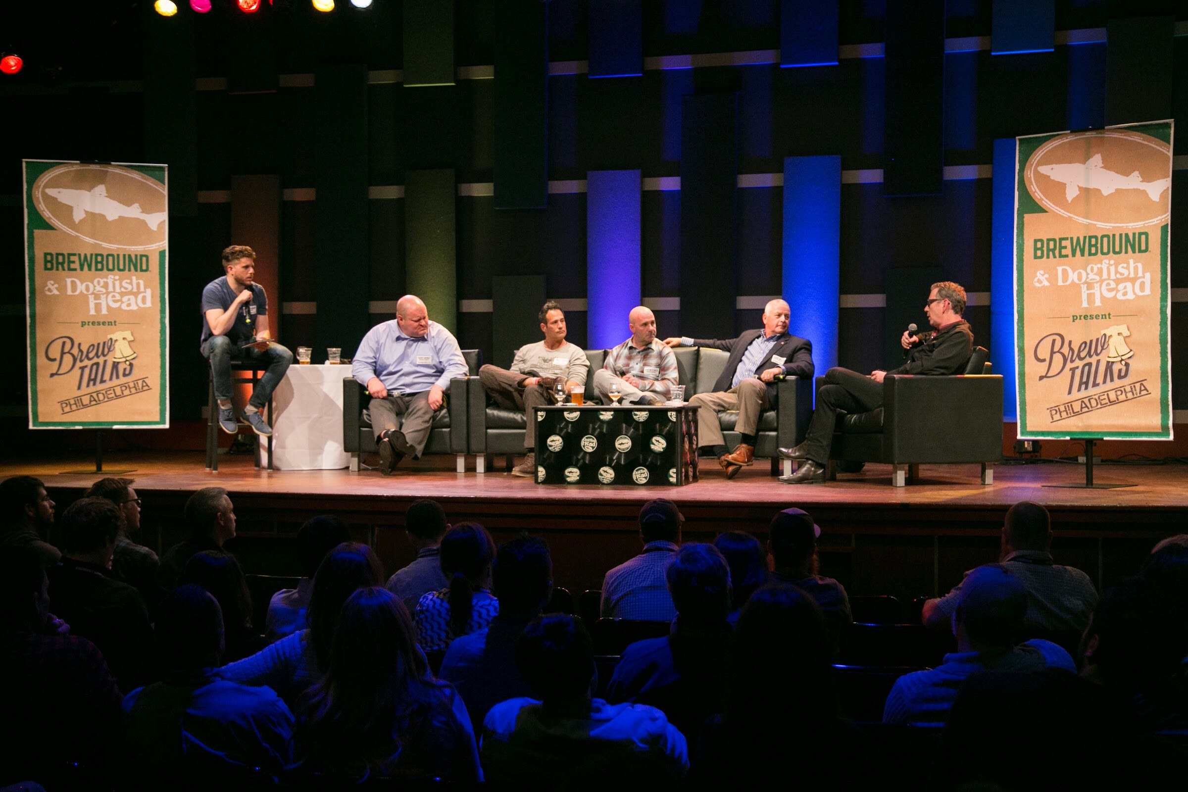 Six men sit across a stage between Brewbound and Dogfish Head banners as audience members watch in the foreground.