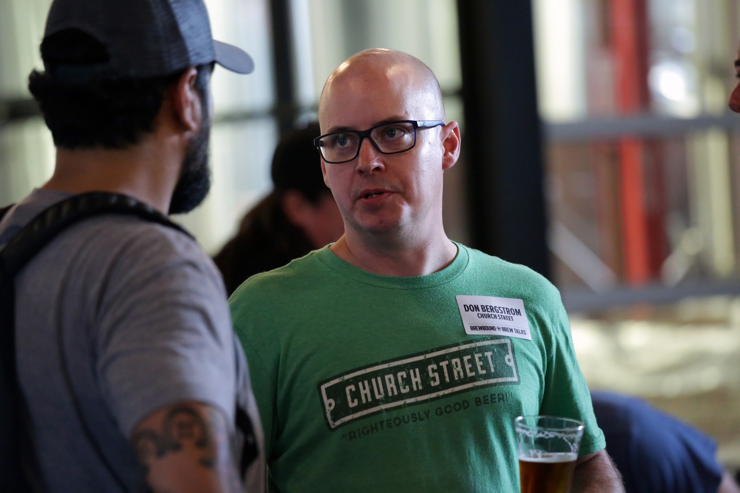 A man wearing a green Church Street shirt and glasses faces a man in a cap.