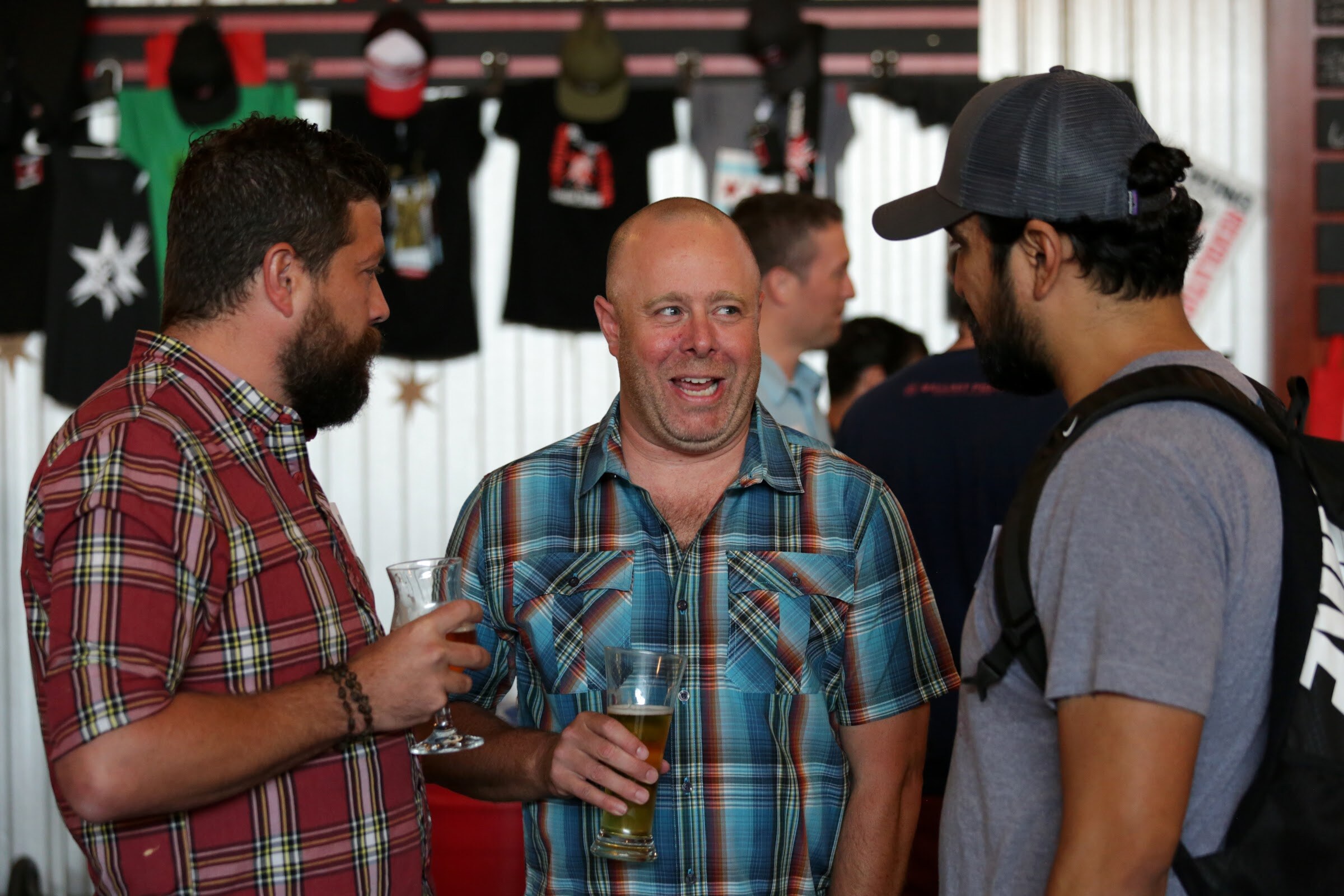 Three men stand together, with the smiling man in the center wearing a blue plaid shirt.
