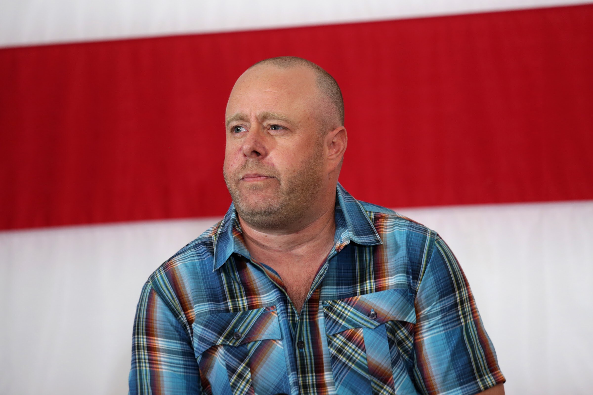 A man in a blue plaid shirt looks to the side in front of red and white flag stripes.