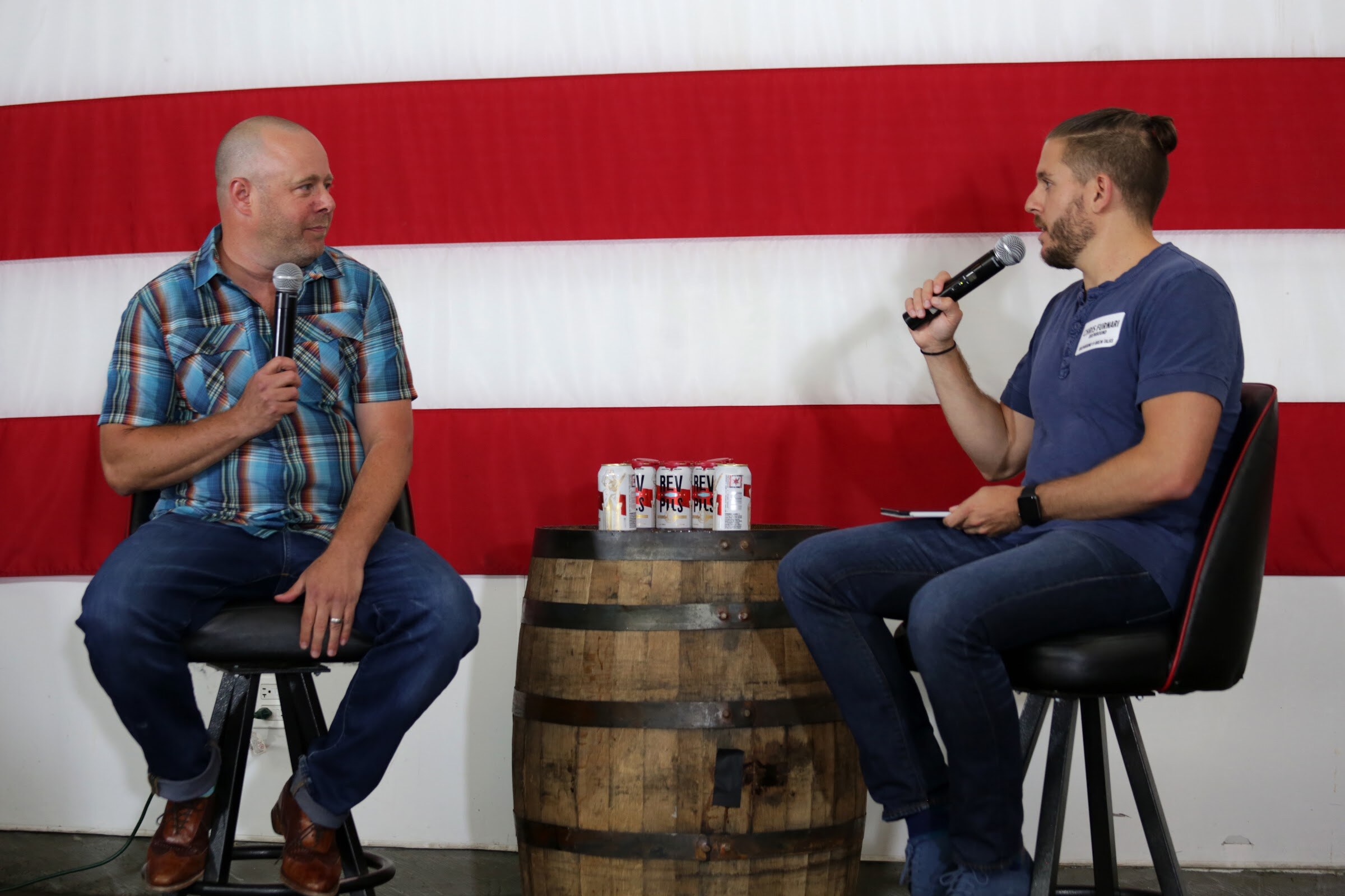 Two men sit on stools facing one another, with beer cans on a barrel between them.