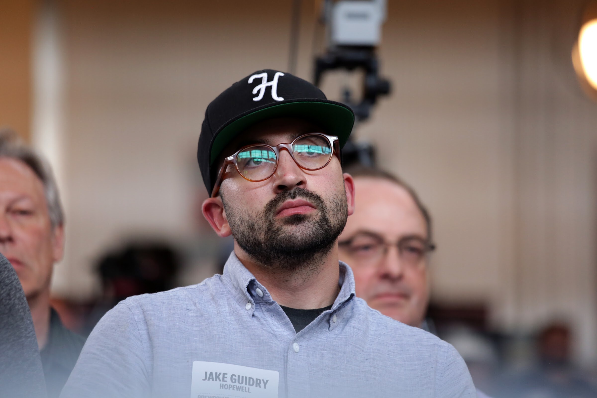 A seated man in a black cap, glasses and light blue shirt looks toward the stage.