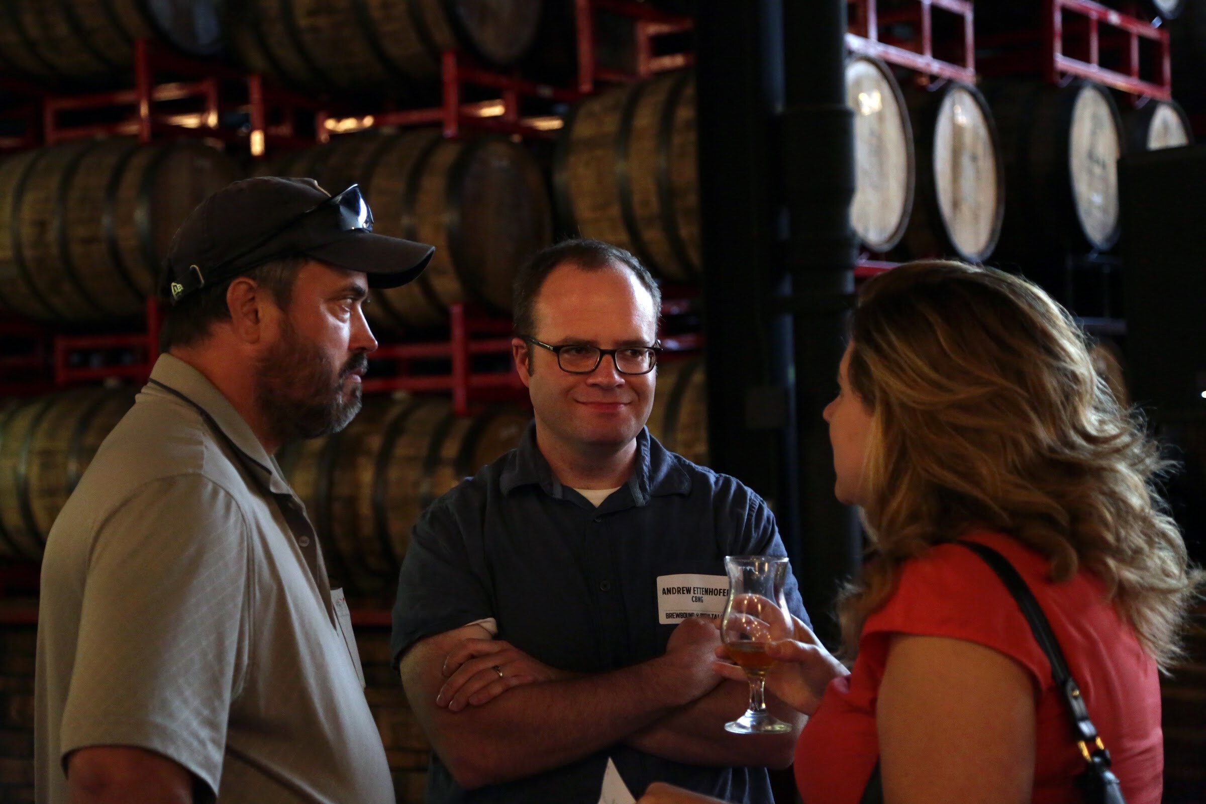 Two men and a woman holding a stemmed glass stand in front of stacked wooden barrels.
