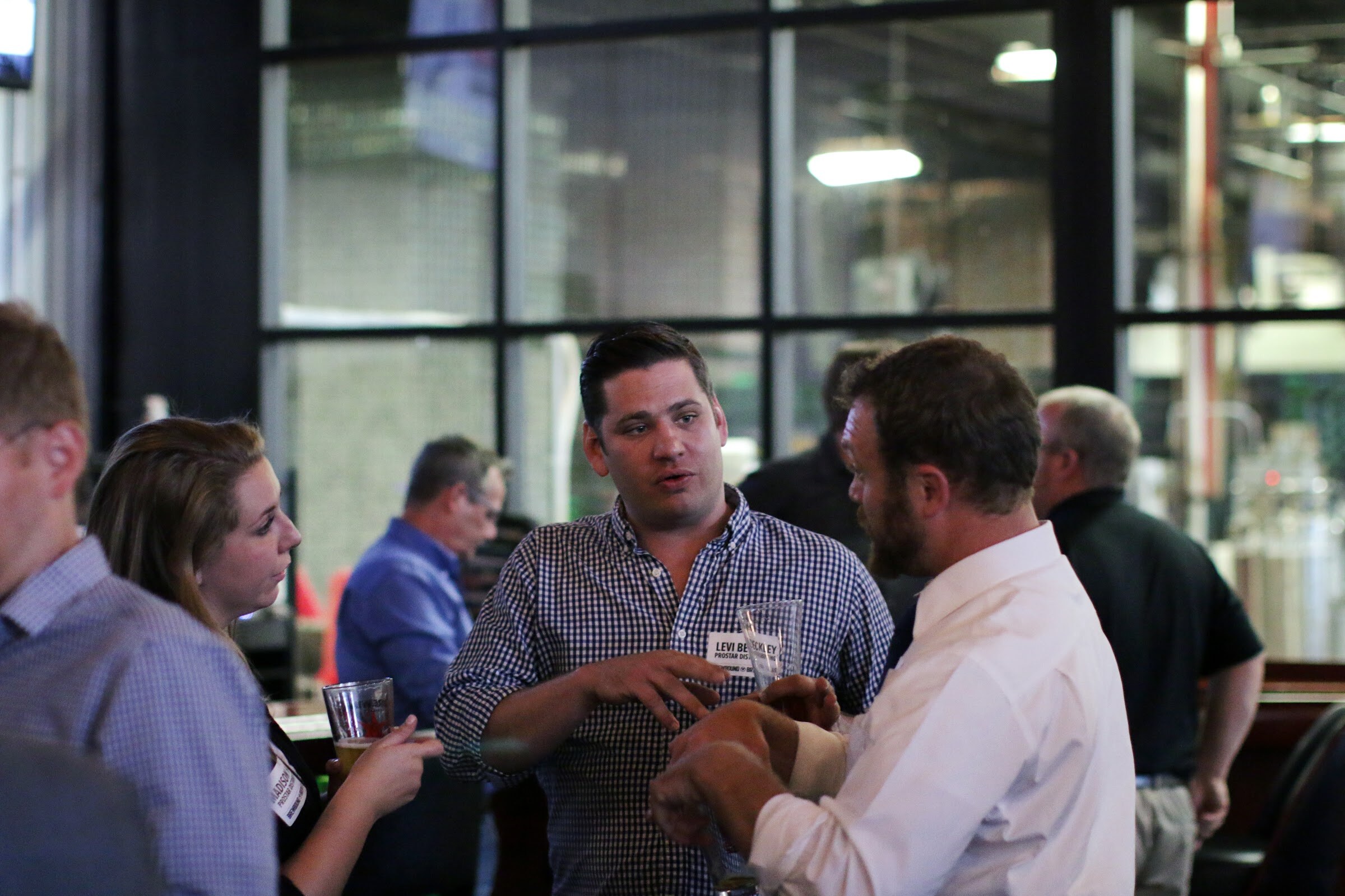 A man in a checked shirt speaks with a woman and a man in a white shirt, all holding drinks.