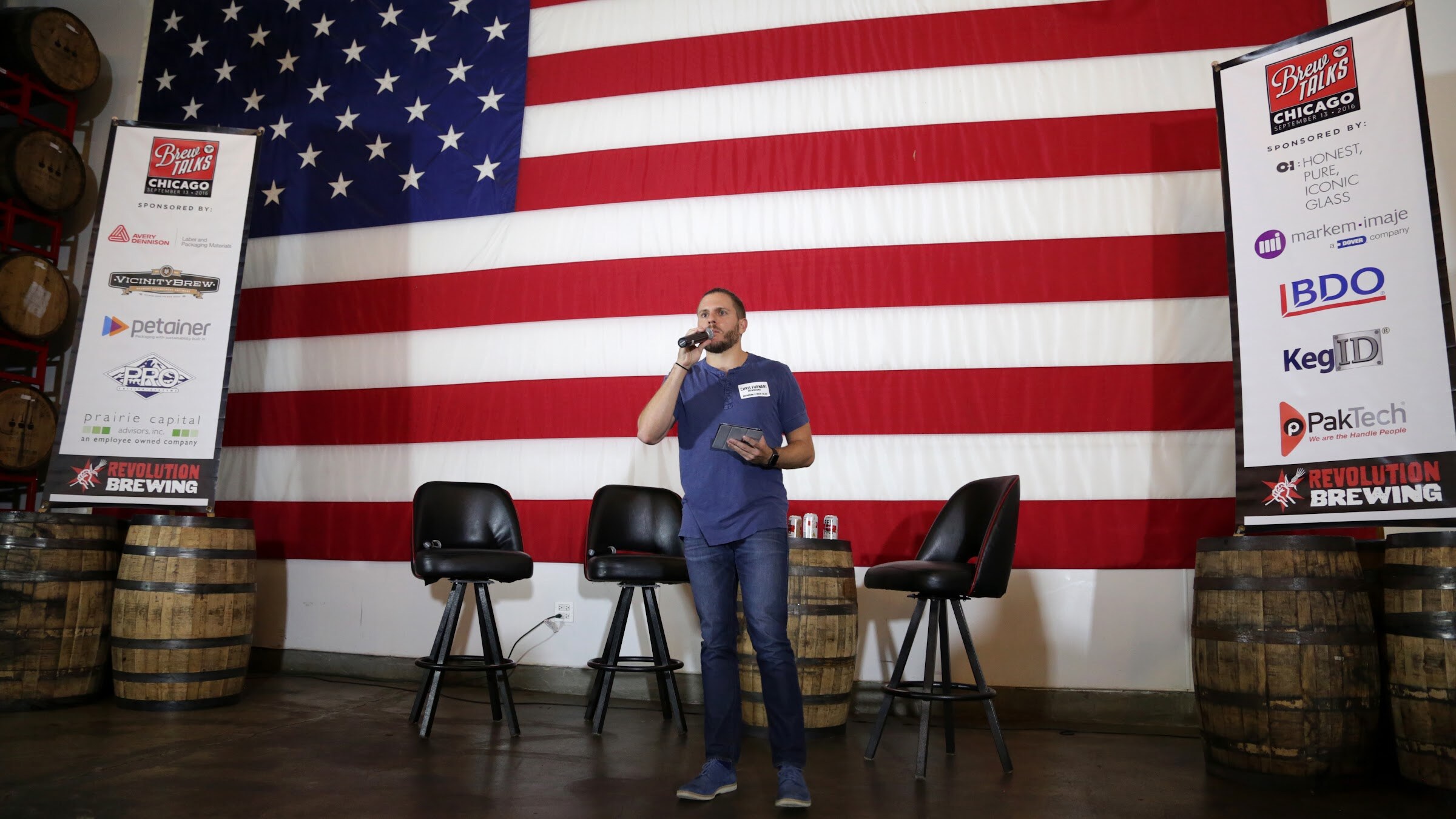 A man with a microphone and tablet stands before an American flag between sponsor banners, stools and barrels.