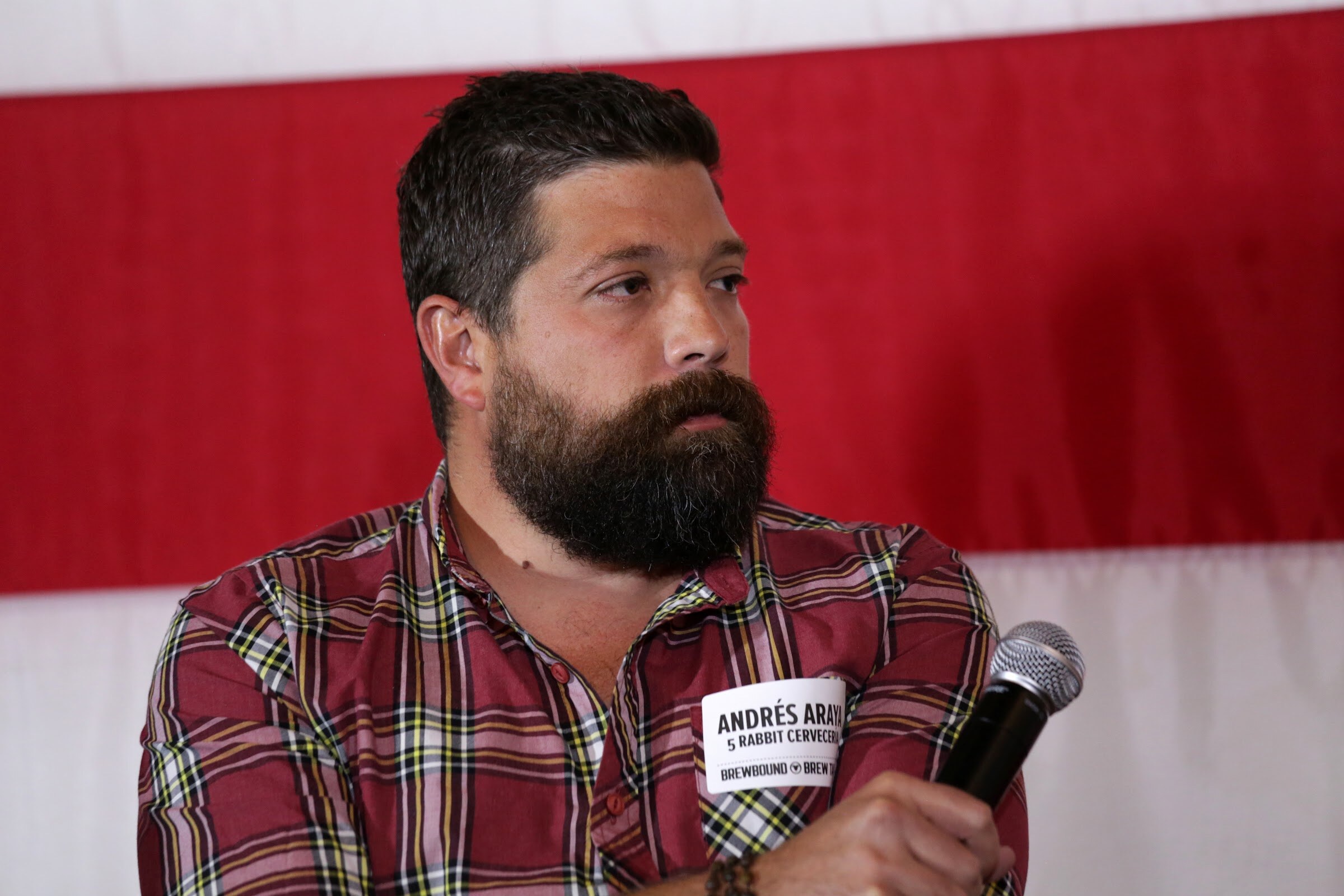 A bearded man in a red plaid shirt holds a microphone in front of red and white flag stripes.