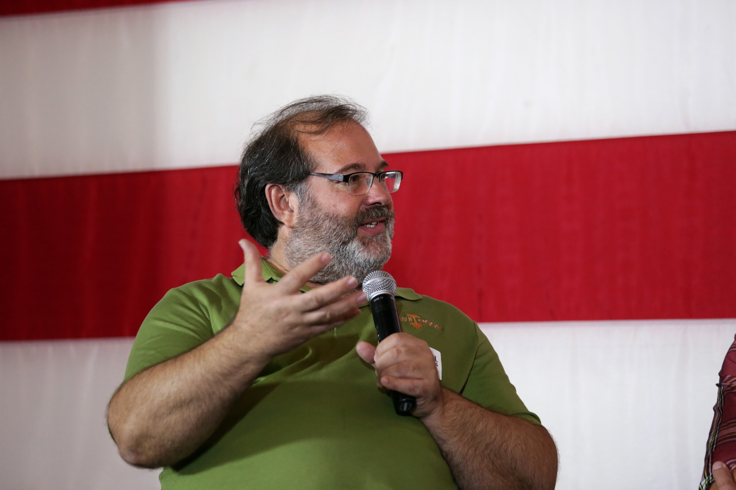 A man in a green polo and glasses raises an open hand while holding a microphone.