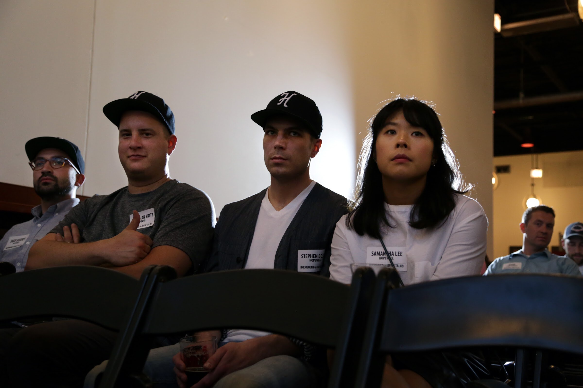 Four audience members sit together, including three wearing black caps, with more guests behind them.