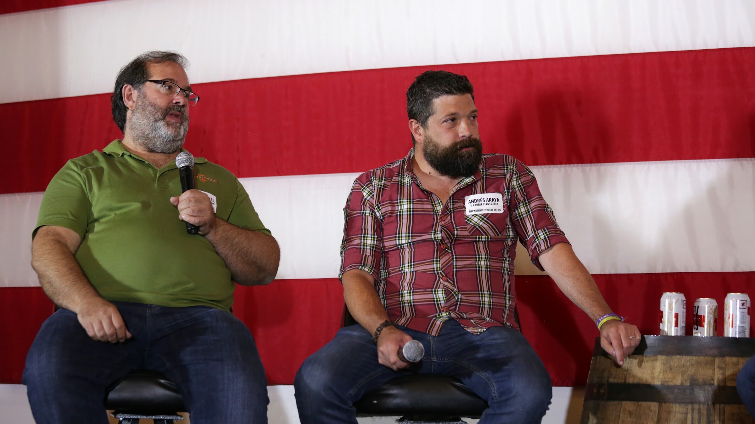 Men in green and red plaid shirts sit before a large flag, with beer cans on a barrel to the right.