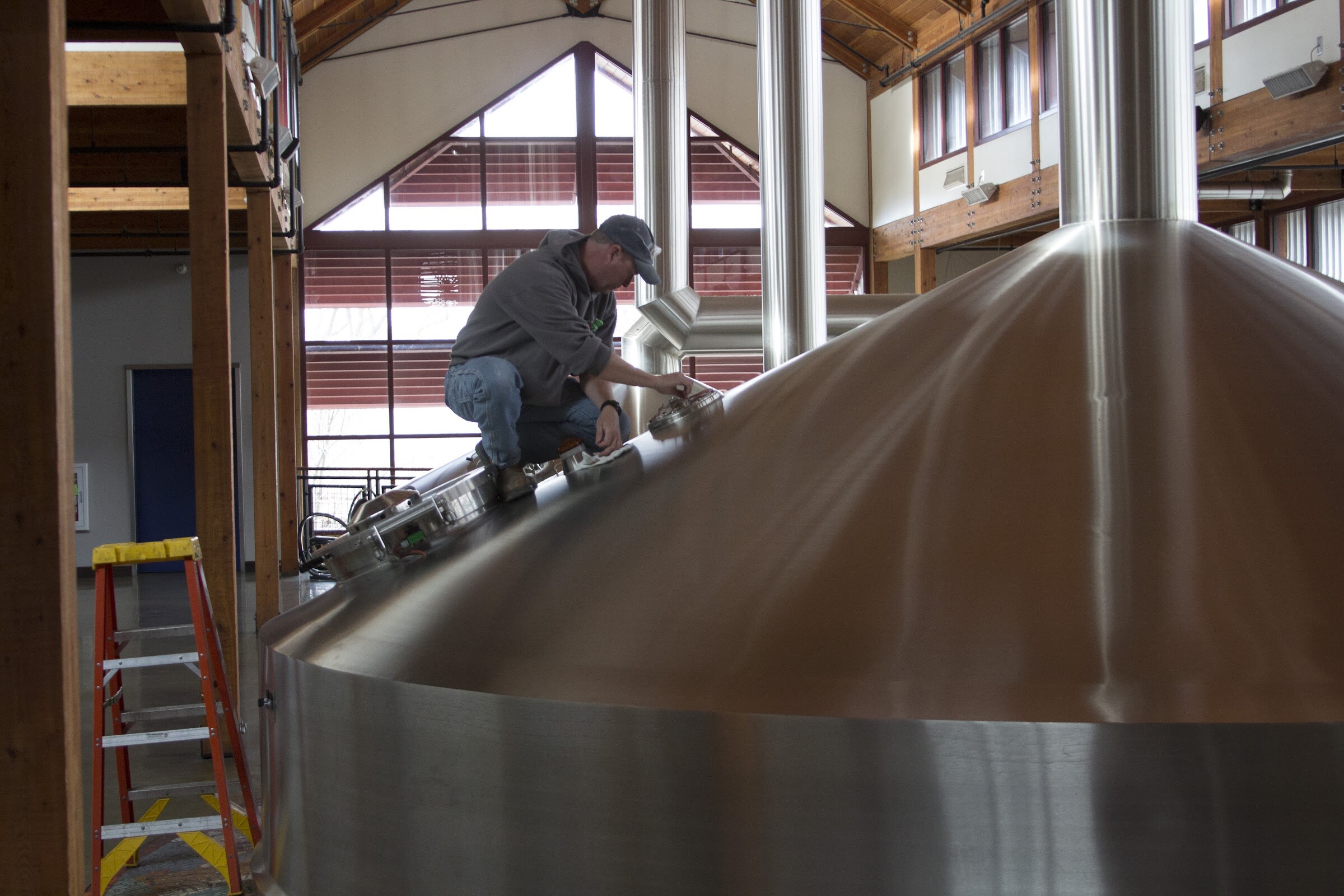 A person crouches over a hatch on a large stainless steel brewing vessel beneath tall windows.