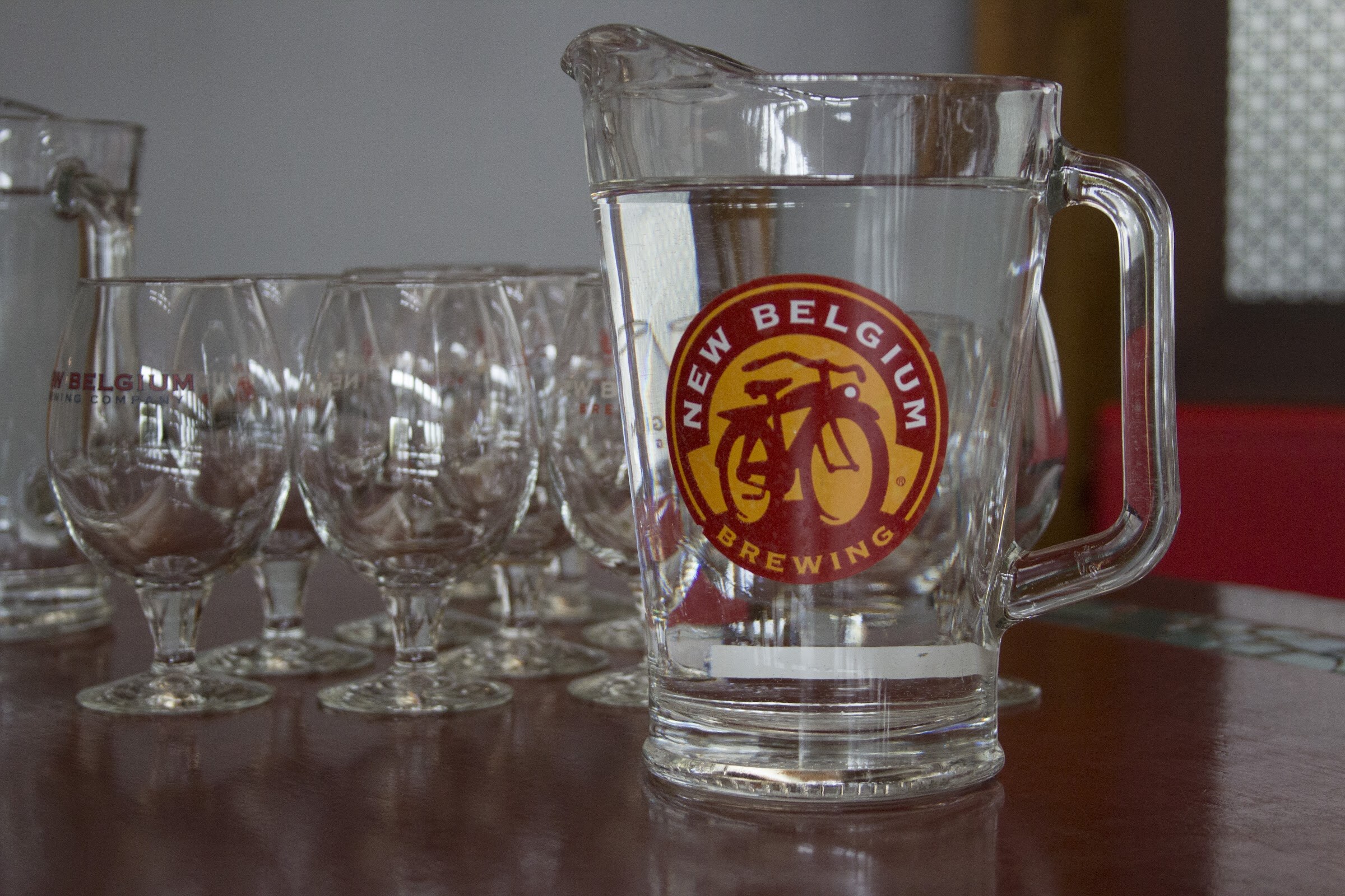 A water pitcher with a New Belgium logo stands beside stemmed glasses on a wooden table.