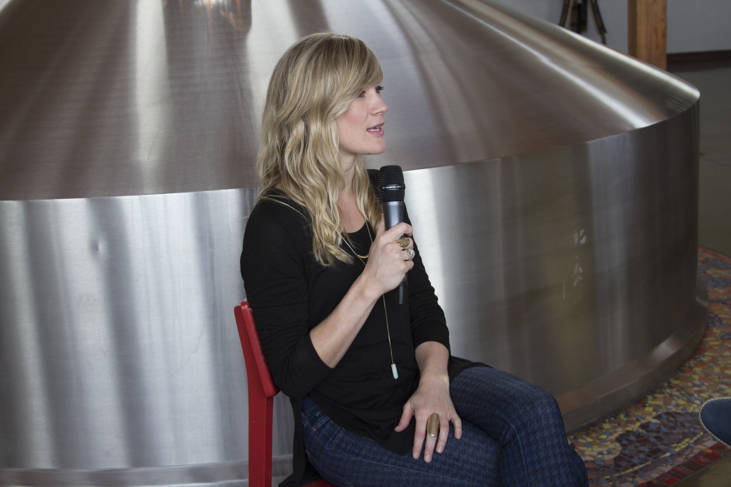 A woman in a black top sits on a red chair holding a microphone beside a large stainless steel vessel.