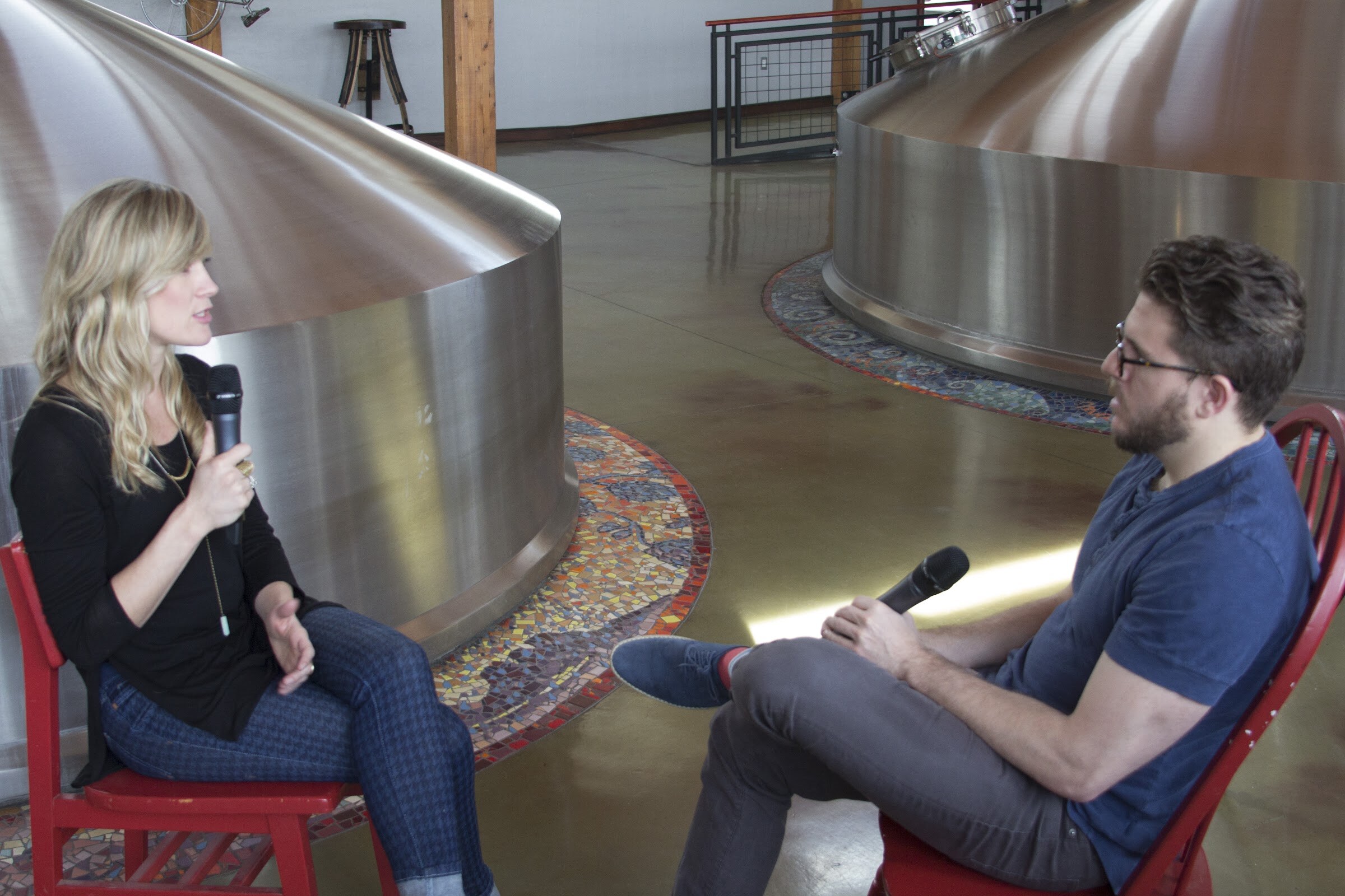 Two people sit on red chairs holding microphones between stainless steel brewing vessels.