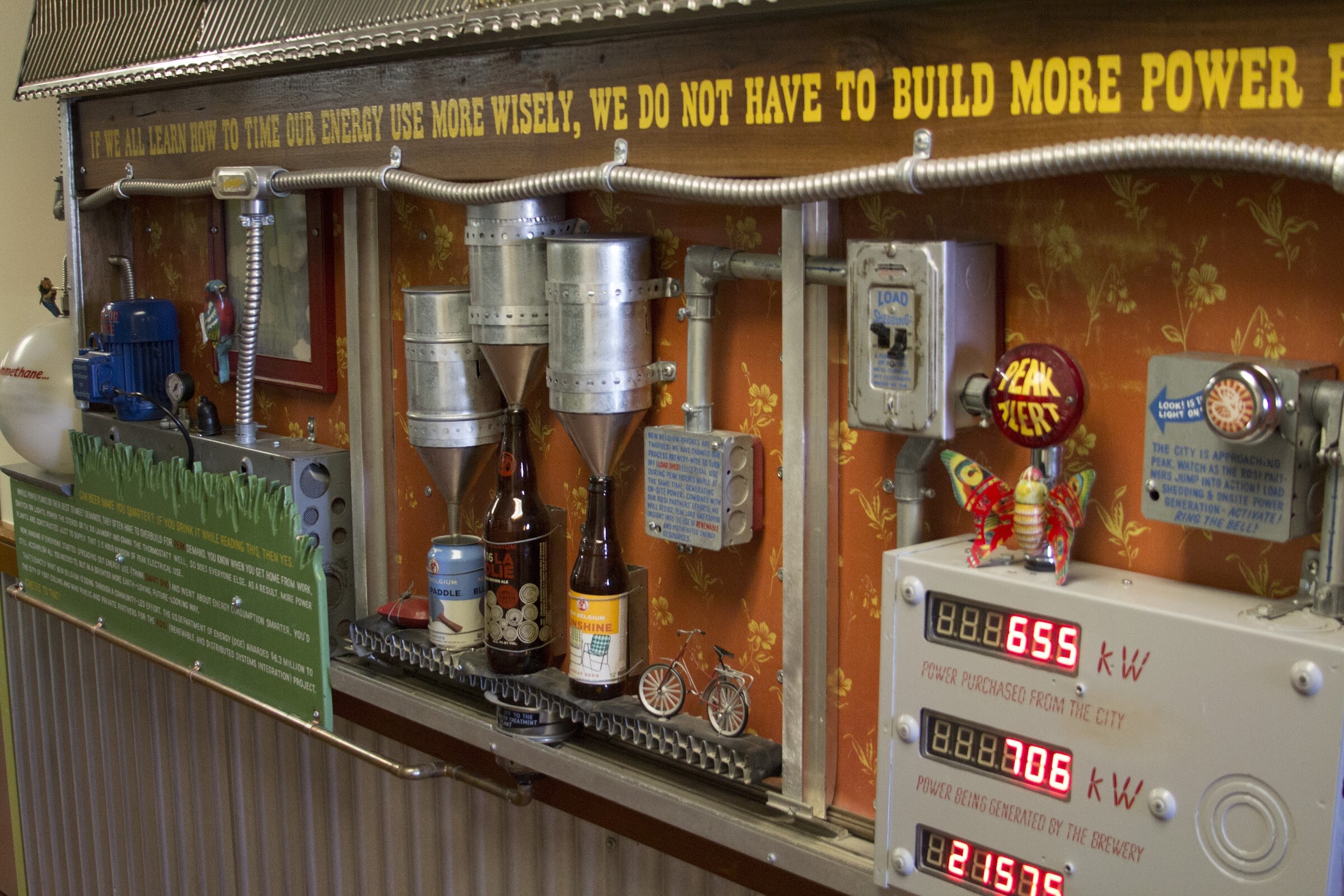 A wall display combines pipes, beer bottles, explanatory signs and illuminated kilowatt readings.