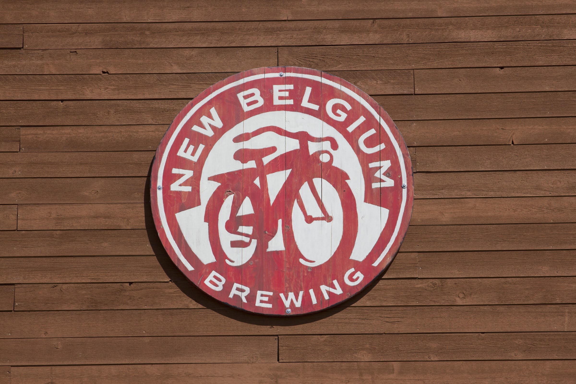 A round red and white New Belgium Brewing bicycle sign hangs on a brown wooden wall.