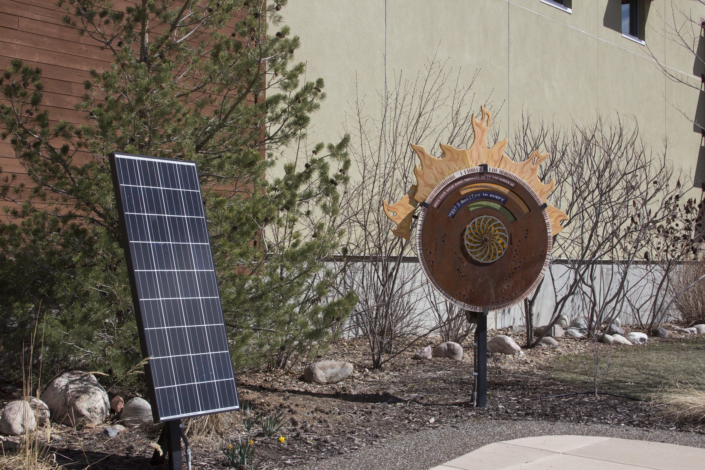 An upright solar panel stands beside a circular sun-shaped display amid shrubs beside a building.