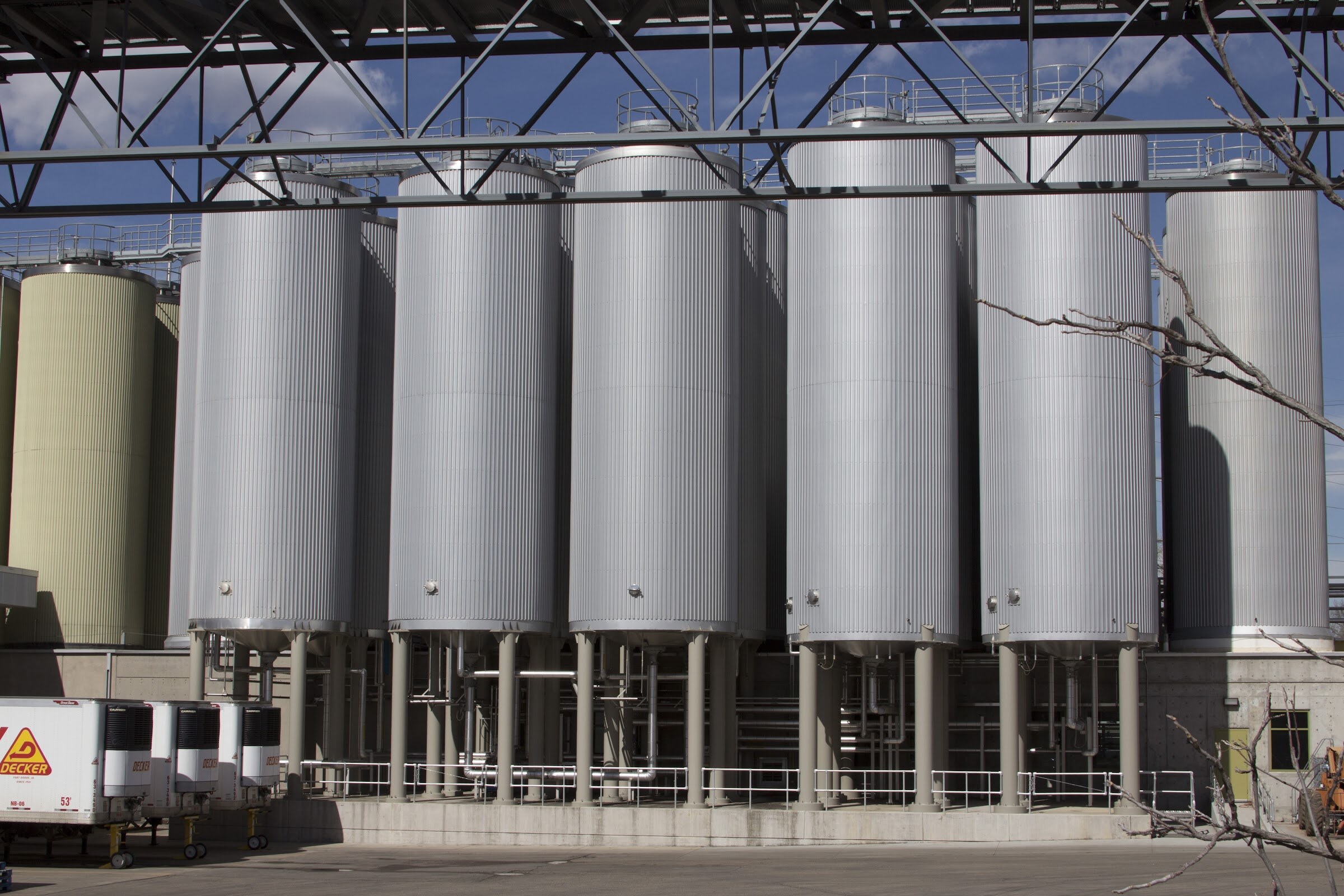 Tall silver cylindrical tanks stand beneath a metal structure outside an industrial building.