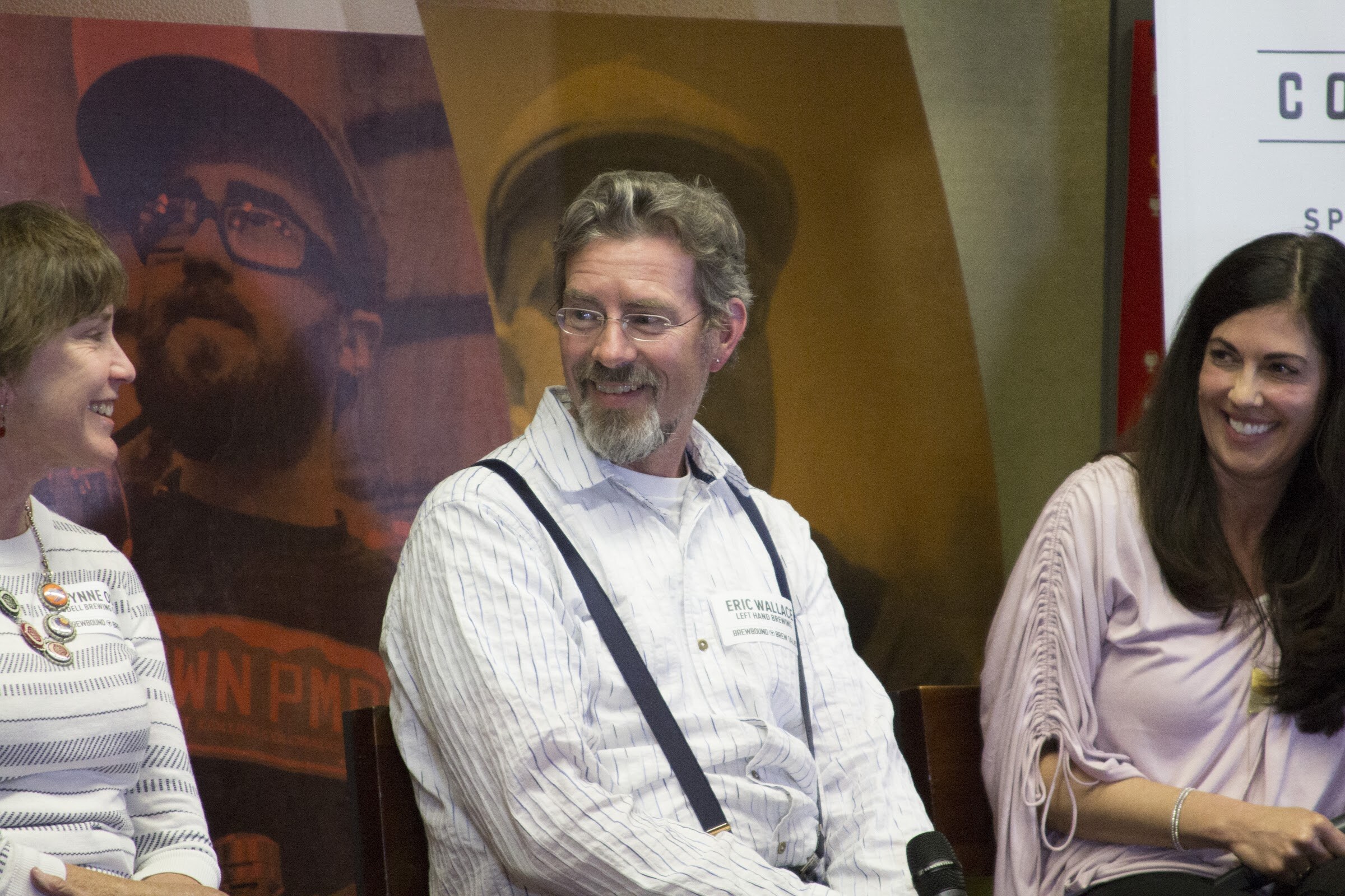 Three seated panelists smile at one another in front of a beer-themed backdrop.