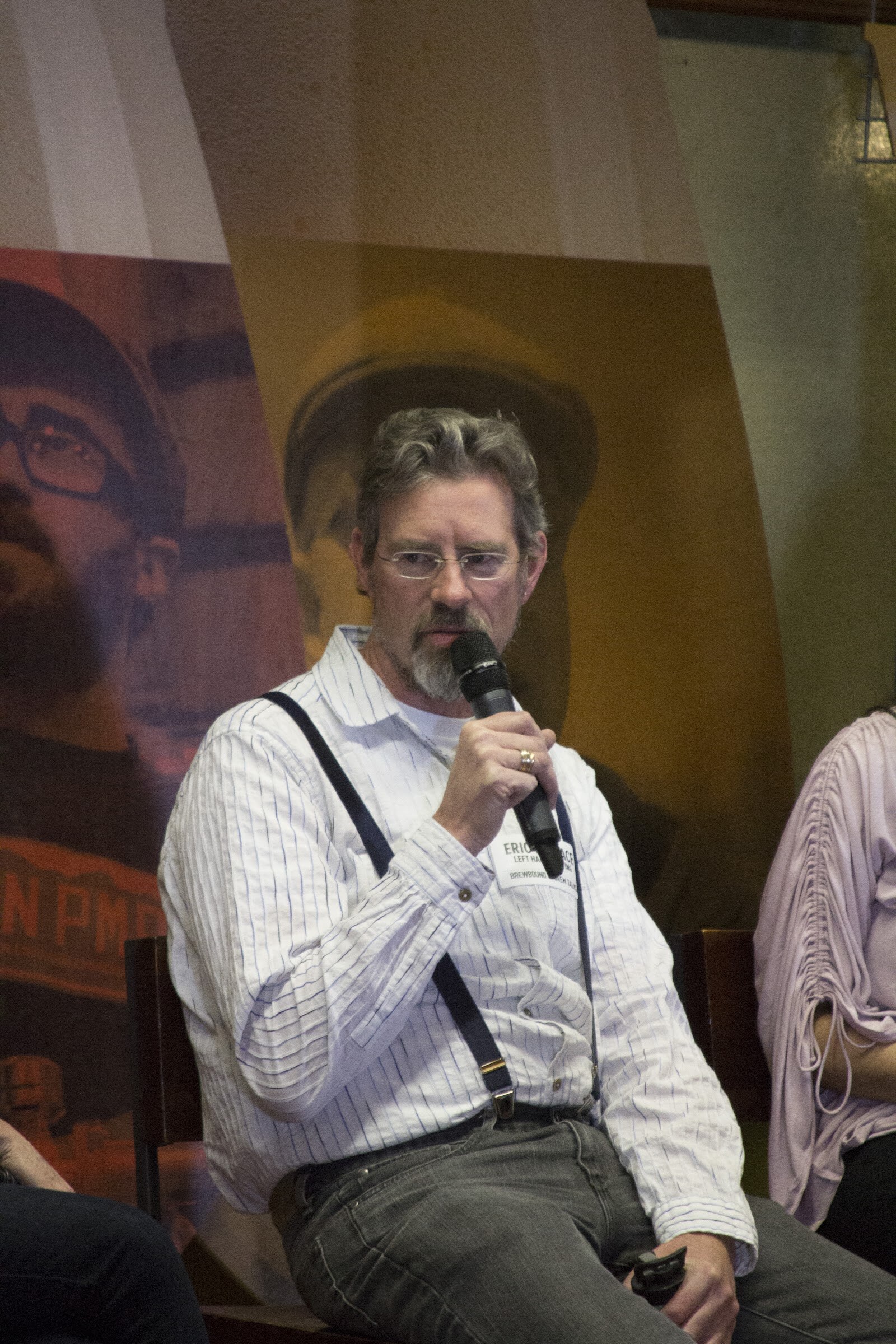 A seated man wearing glasses, a striped shirt and suspenders holds a microphone near his mouth.