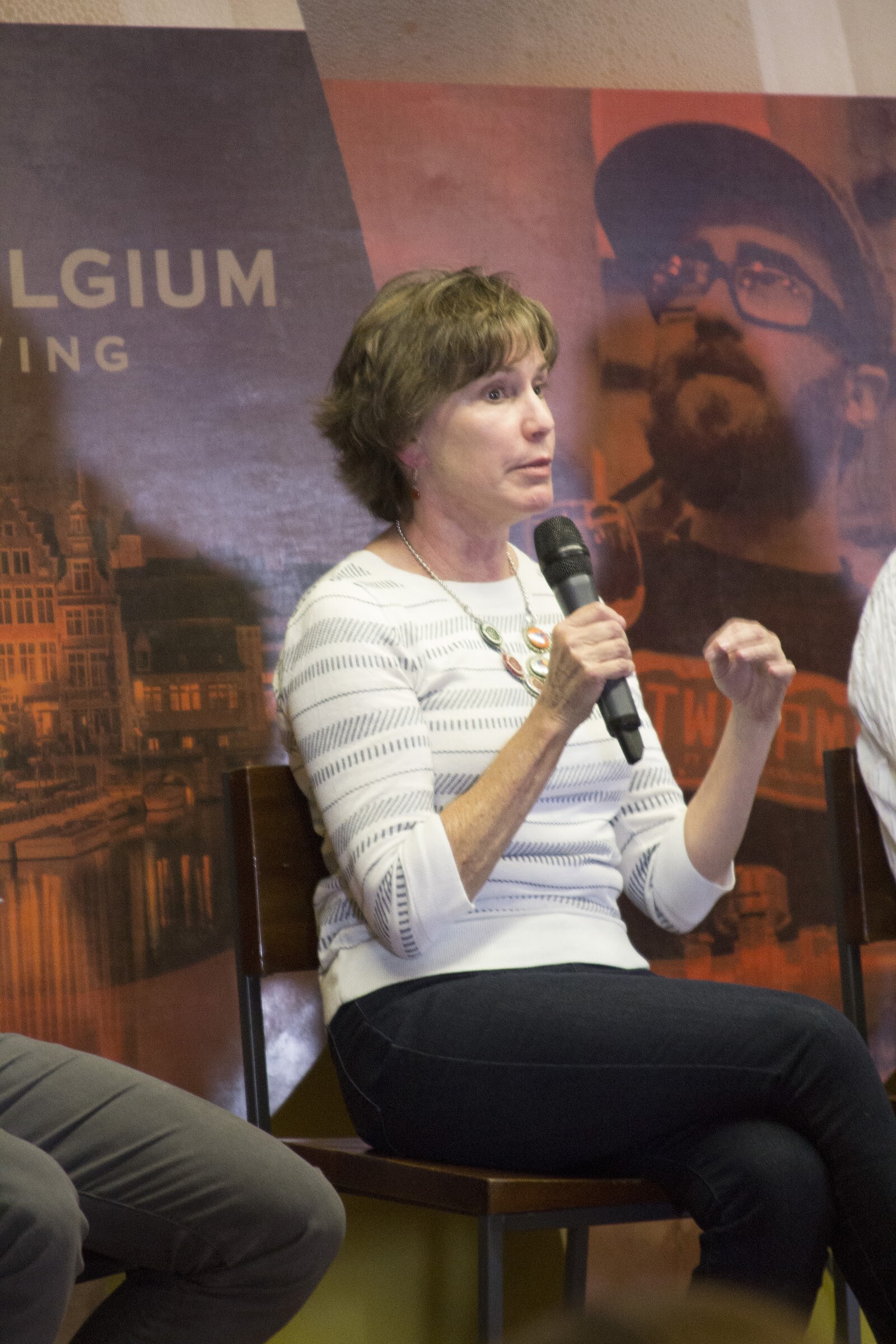 A woman in a white patterned top holds a microphone and raises her free hand beside a beer-themed backdrop.