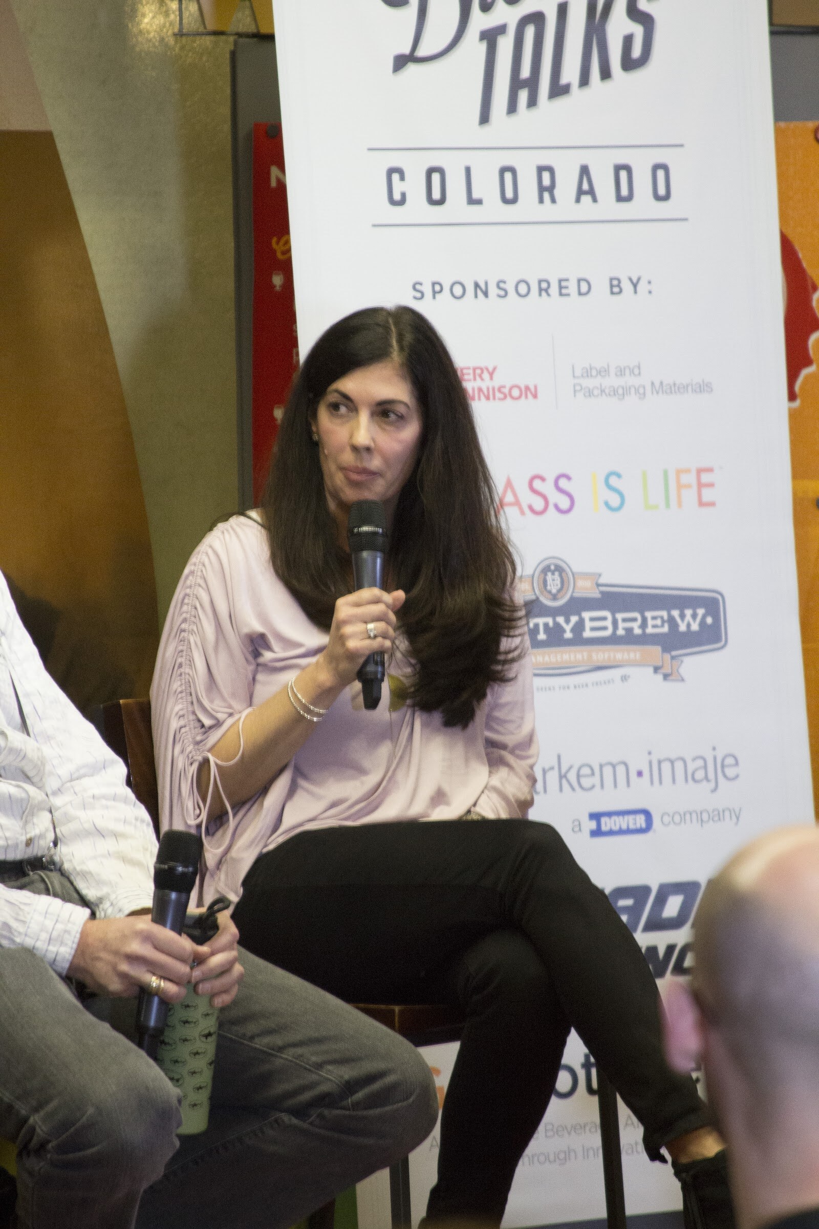 A woman in a pale blouse sits with crossed legs in front of a Brew Talks Colorado sponsor banner.