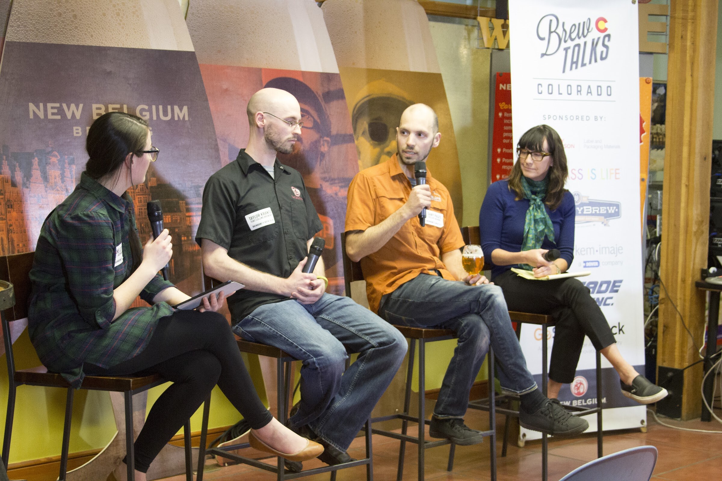 Four people sit on tall chairs as a man in an orange shirt speaks into a microphone while holding a glass of beer.