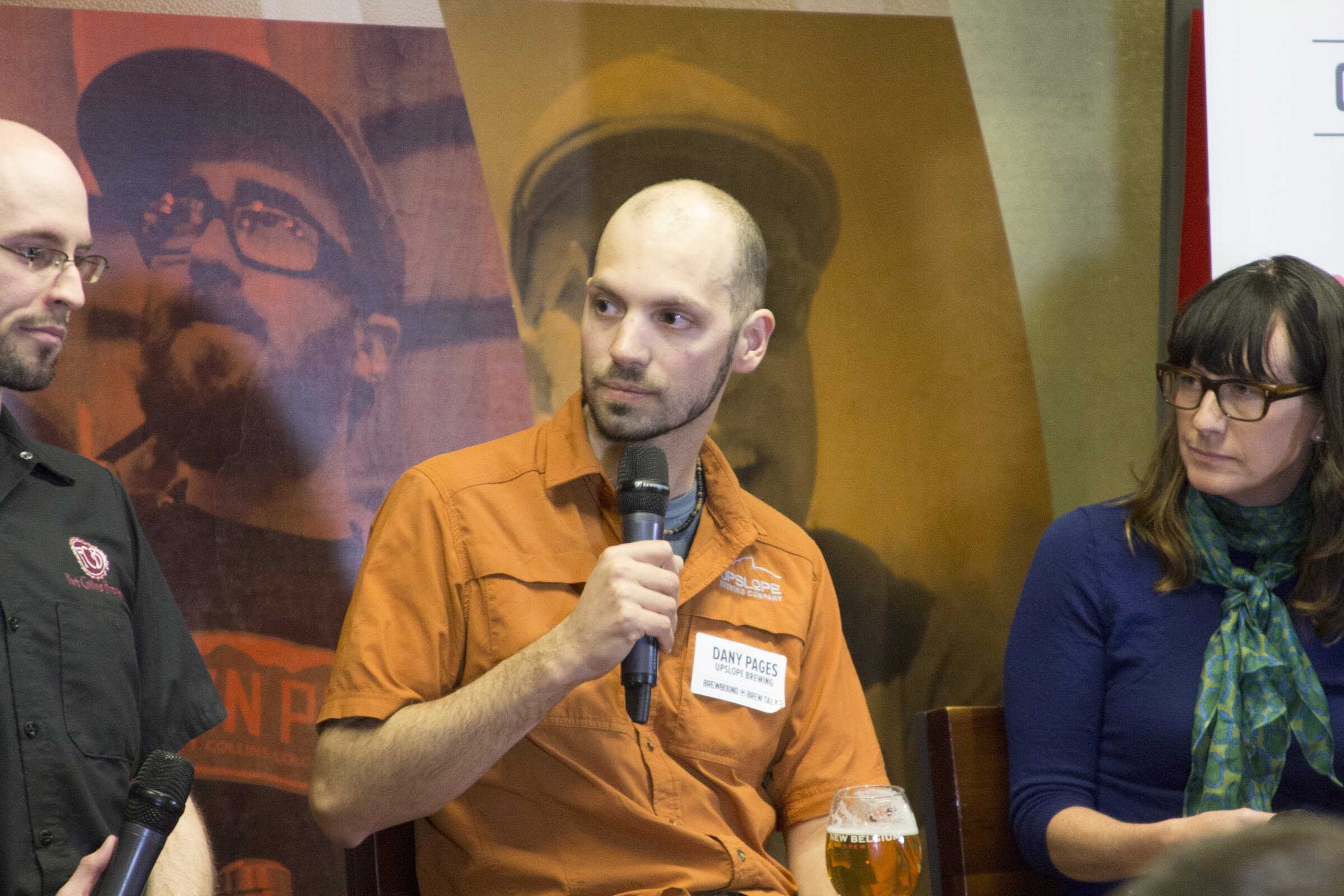 A man in an orange shirt holds a microphone between two seated panelists in front of a beer-themed backdrop.