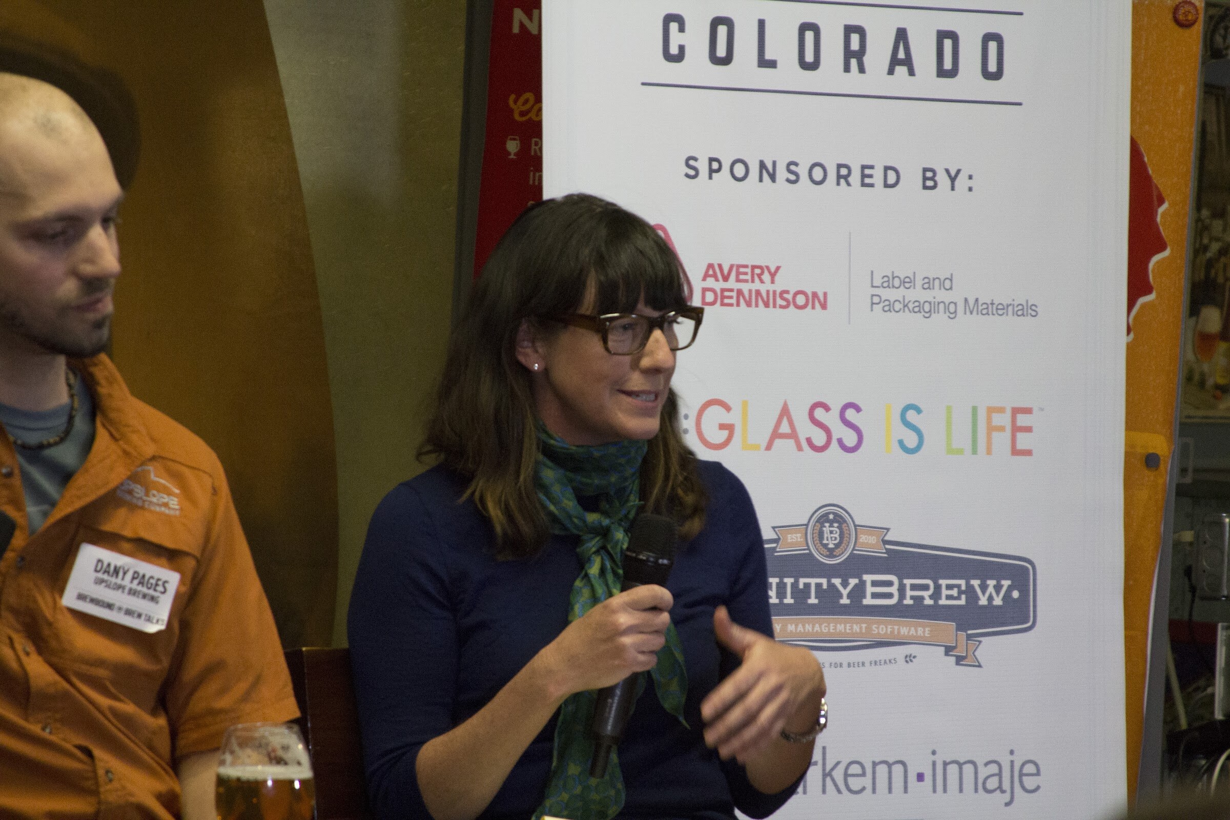 A woman wearing glasses and a green scarf holds a microphone in front of the event's sponsor banner.