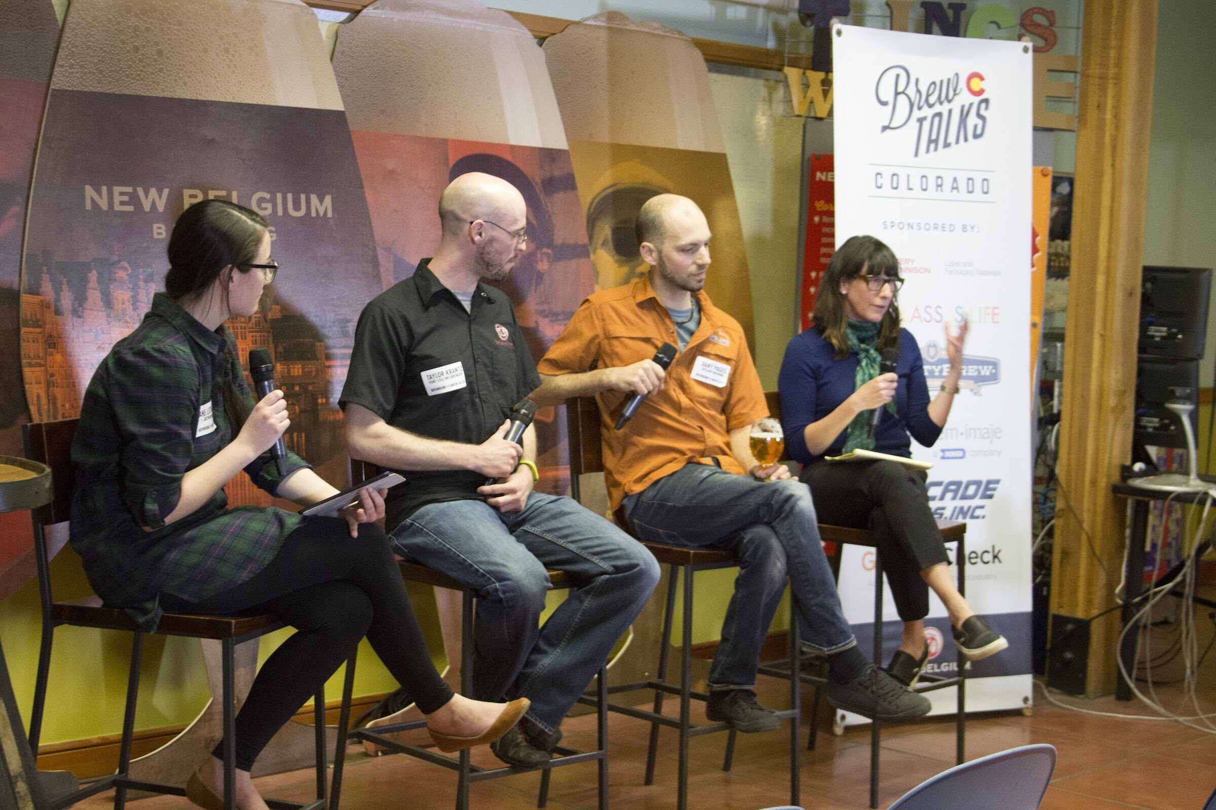 Four seated panel participants face toward a woman in a blue top who gestures with her free hand.