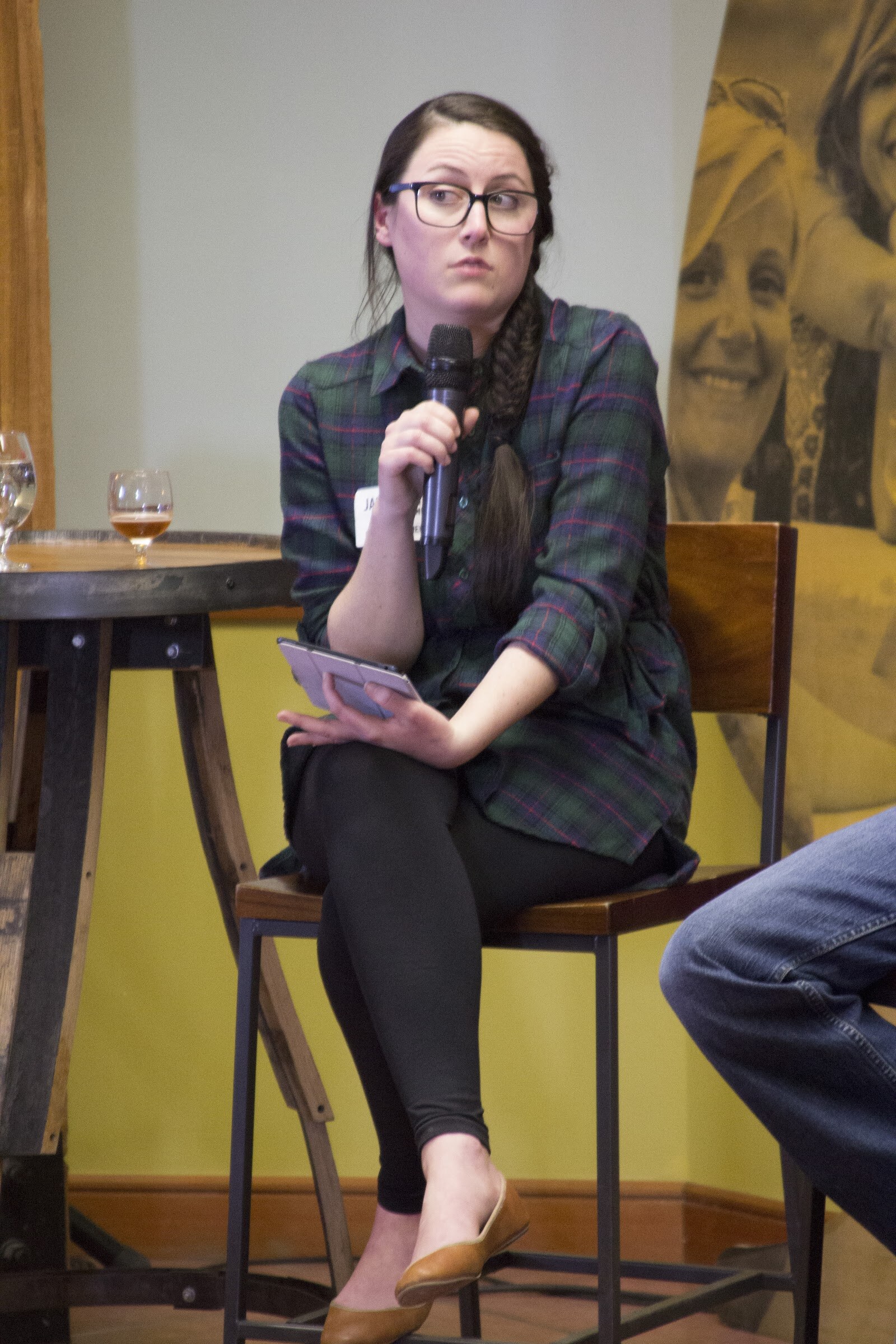 A woman in glasses and a plaid dress sits on a tall chair with a microphone and notecards.