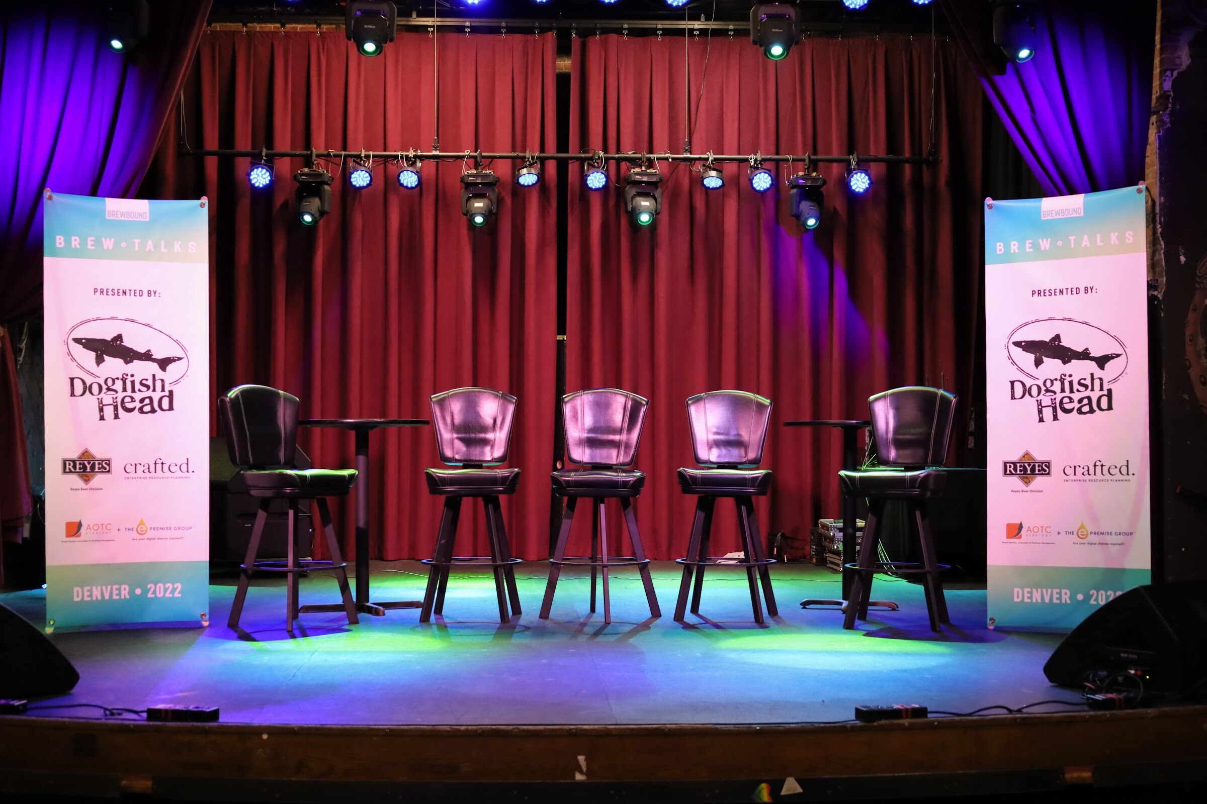 Five empty black leather swivel bar stools on a stage in front of a red curtain with blue and green stage lights, flanked by two tall Brewbound Brew Talks banners reading Presented by Dogfish Head, Reyes, crafted and Denver 2022.