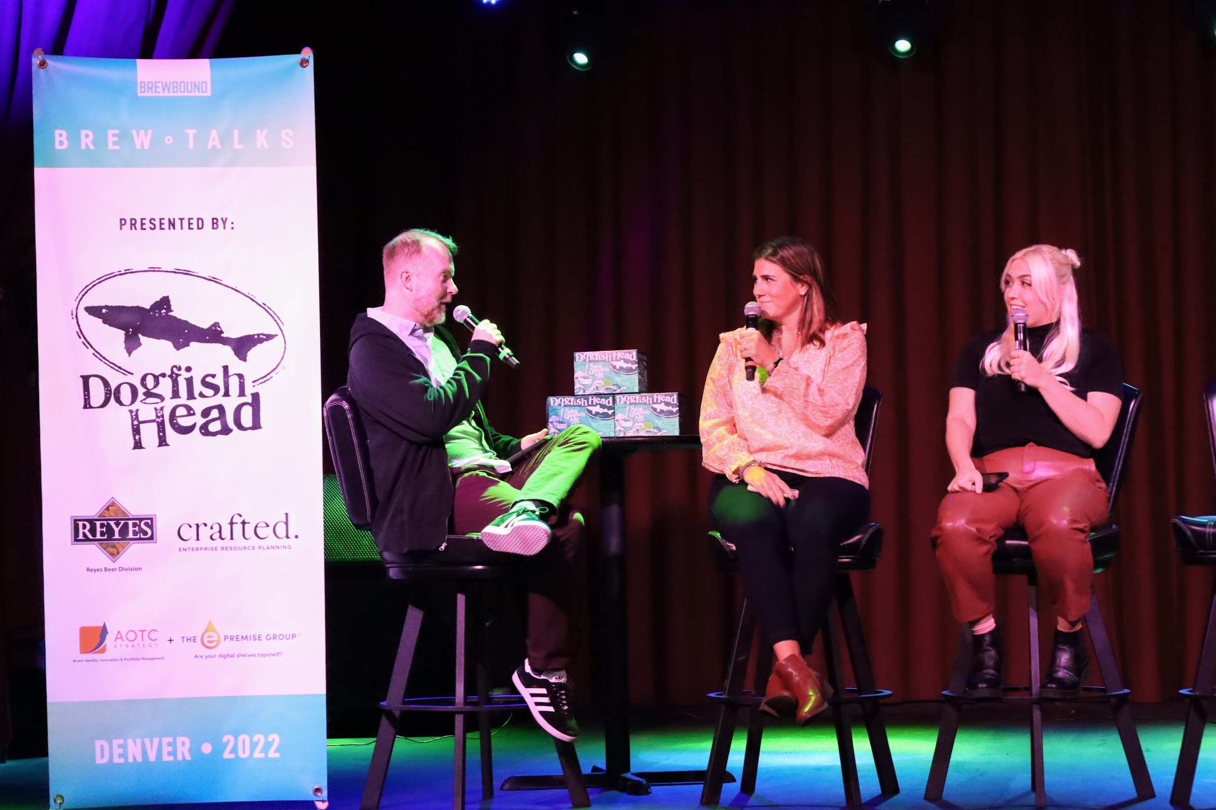 A man in a dark cardigan and bright green pants and two women, one in a peach ruffled blouse and one with white-blonde hair in a black top and rust leather pants, hold microphones on bar stools beside a table with Dogfish Head cartons and a Brew Talks banner.