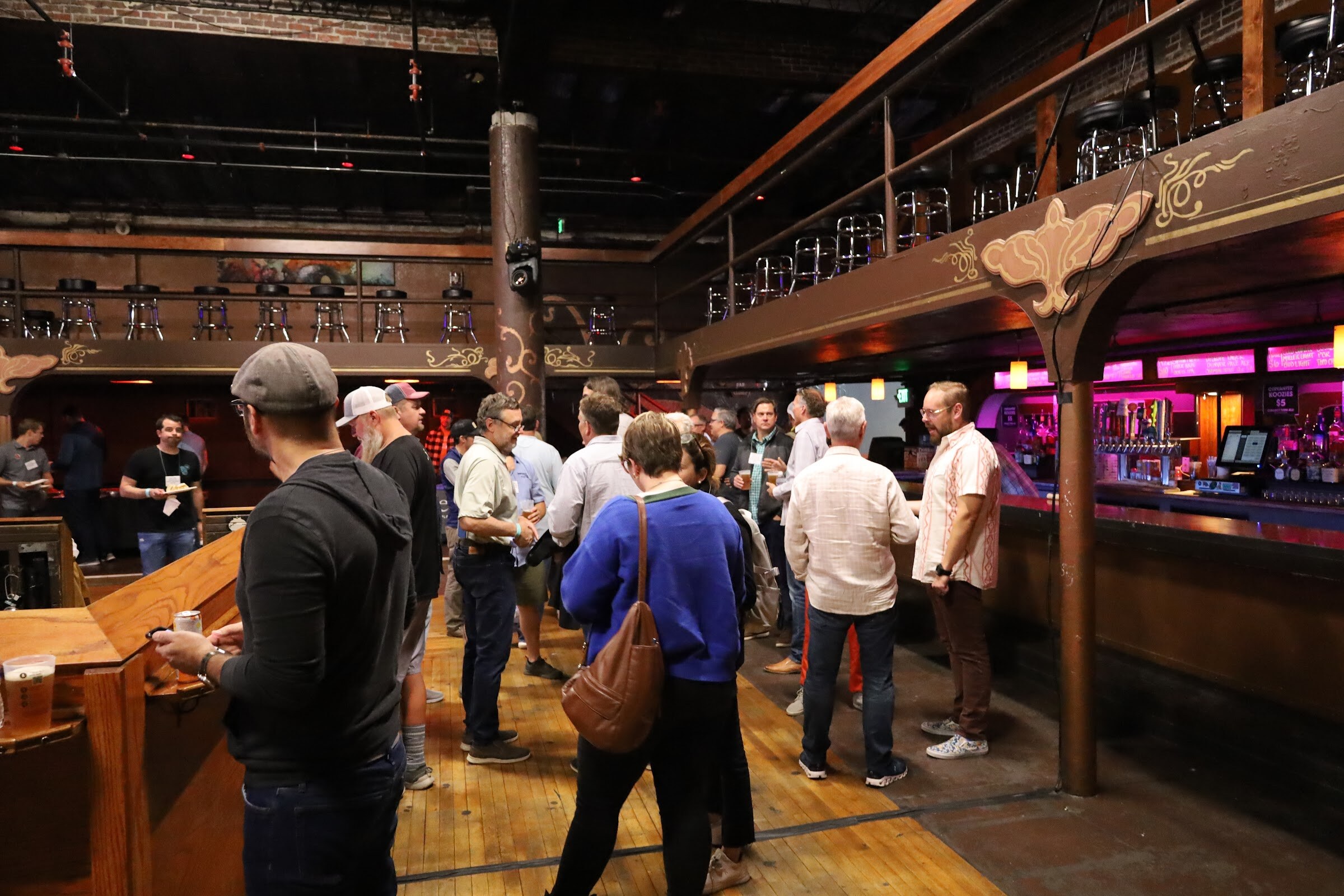 Guests with name badges stand and talk on a wooden floor beside a long pink-lit bar, under a wooden balcony lined with bar stools, with a man in a gray cap and black hooded sweatshirt holding a beer in the foreground.