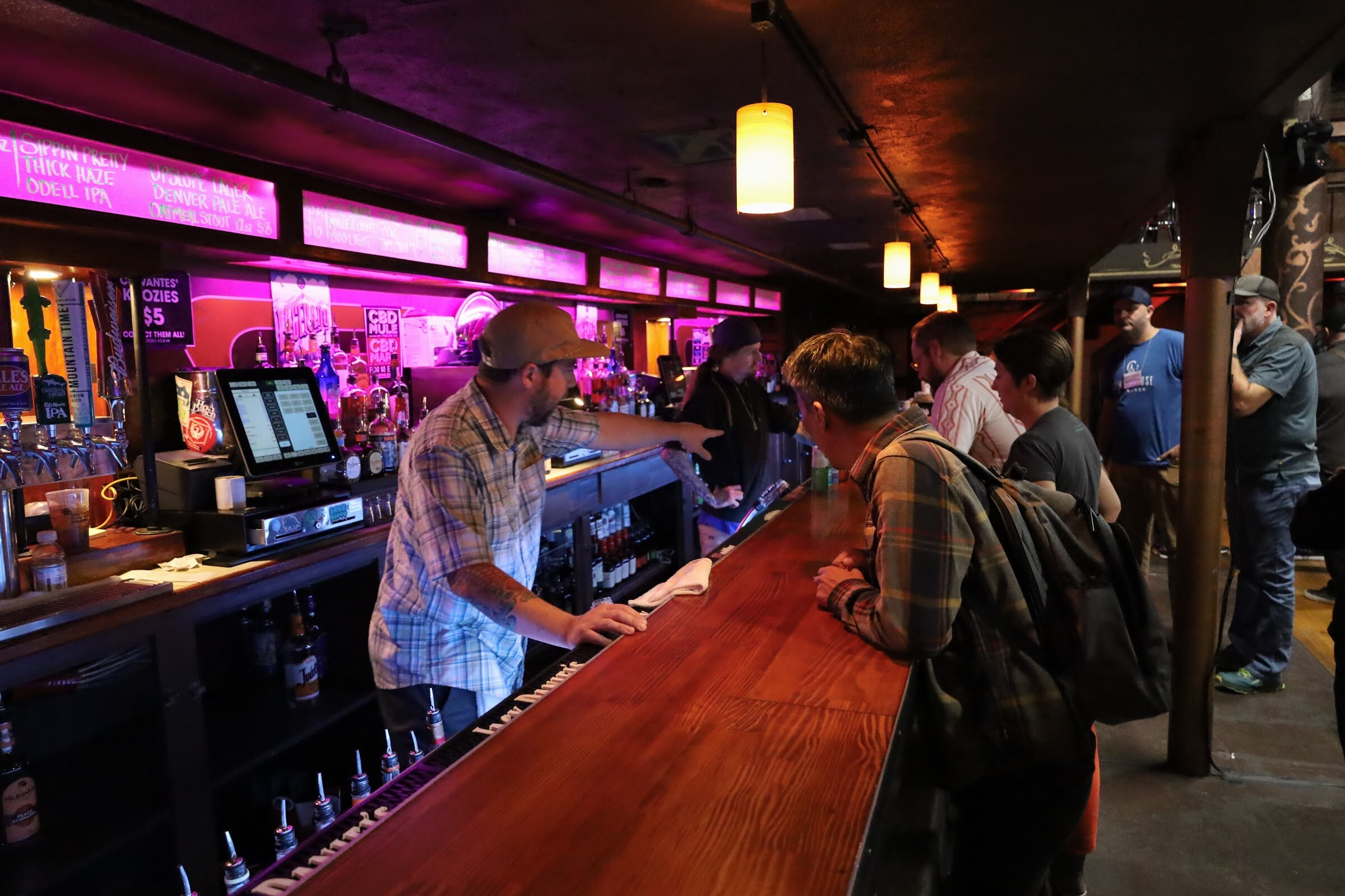 A bartender in a light plaid shirt and tan cap points along a polished wooden bar lit in pink, while a guest in a plaid jacket and backpack leans on the counter, with chalkboard beer menus above and other guests behind.
