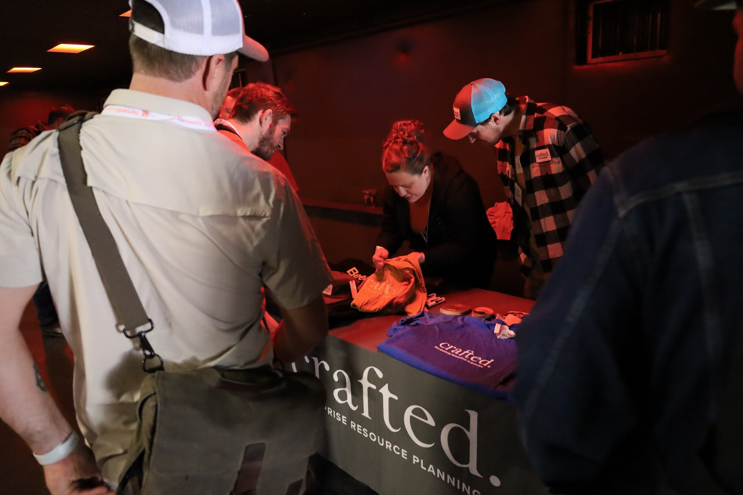 A man in a tan shirt with a messenger bag, a man in a teal and white cap and red plaid shirt and a woman in black lean over a table with a gray crafted. Enterprise Resource Planning cover, folding a yellow shirt beside a purple crafted. shirt.