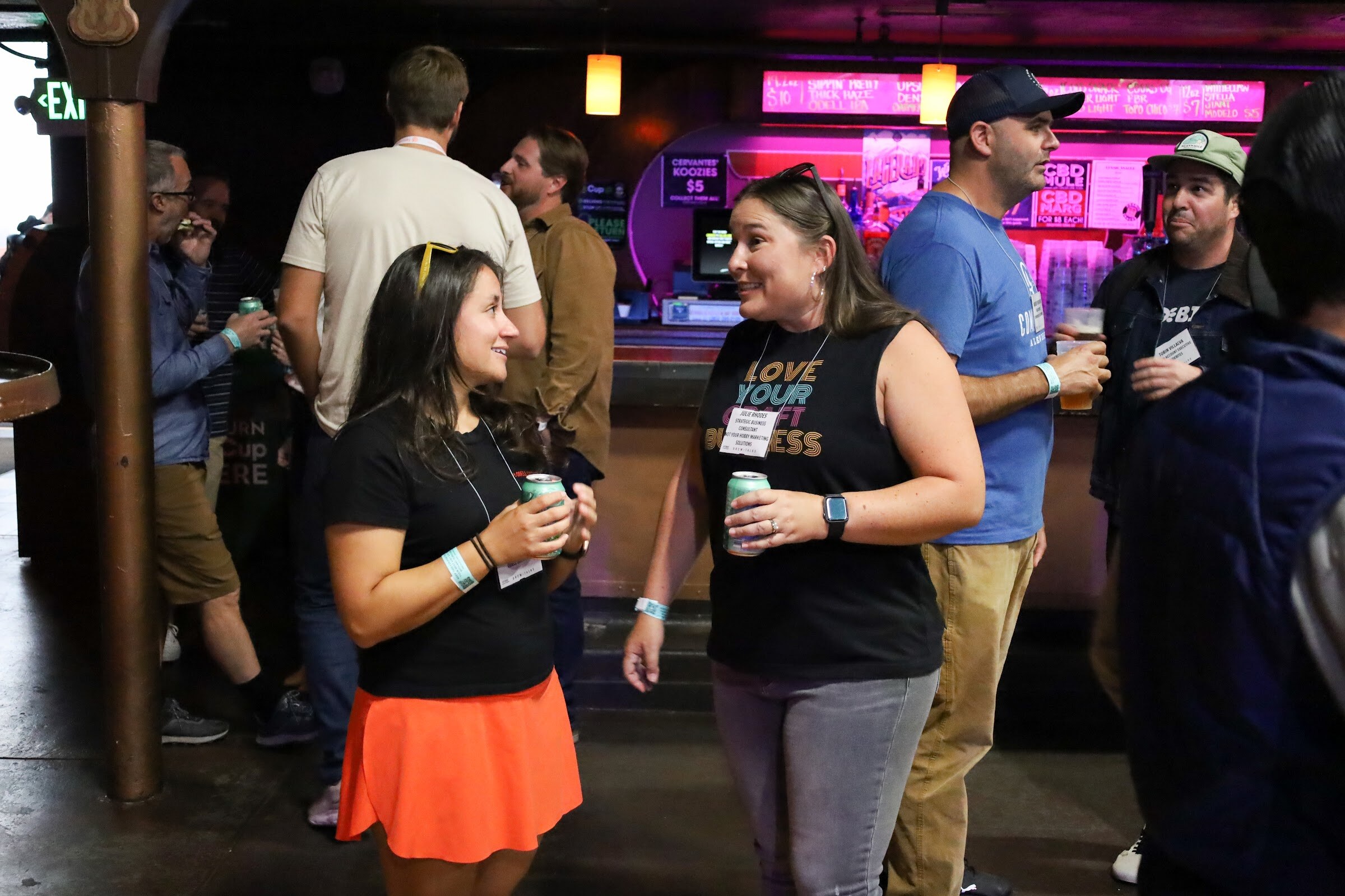 A woman in a black top and orange skirt and a woman in a black Love Your Craft Business tank top hold green cans and smile at each other, with other guests and the pink-lit bar behind them.