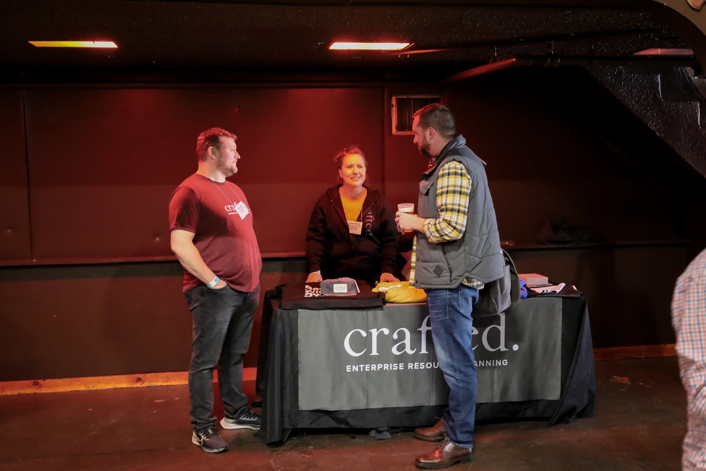 A man in a maroon t-shirt and a smiling woman in a black hoodie stand at a table with a gray crafted. Enterprise Resource Planning cover while a bearded man in a gray vest and plaid shirt holding a cup talks with them.