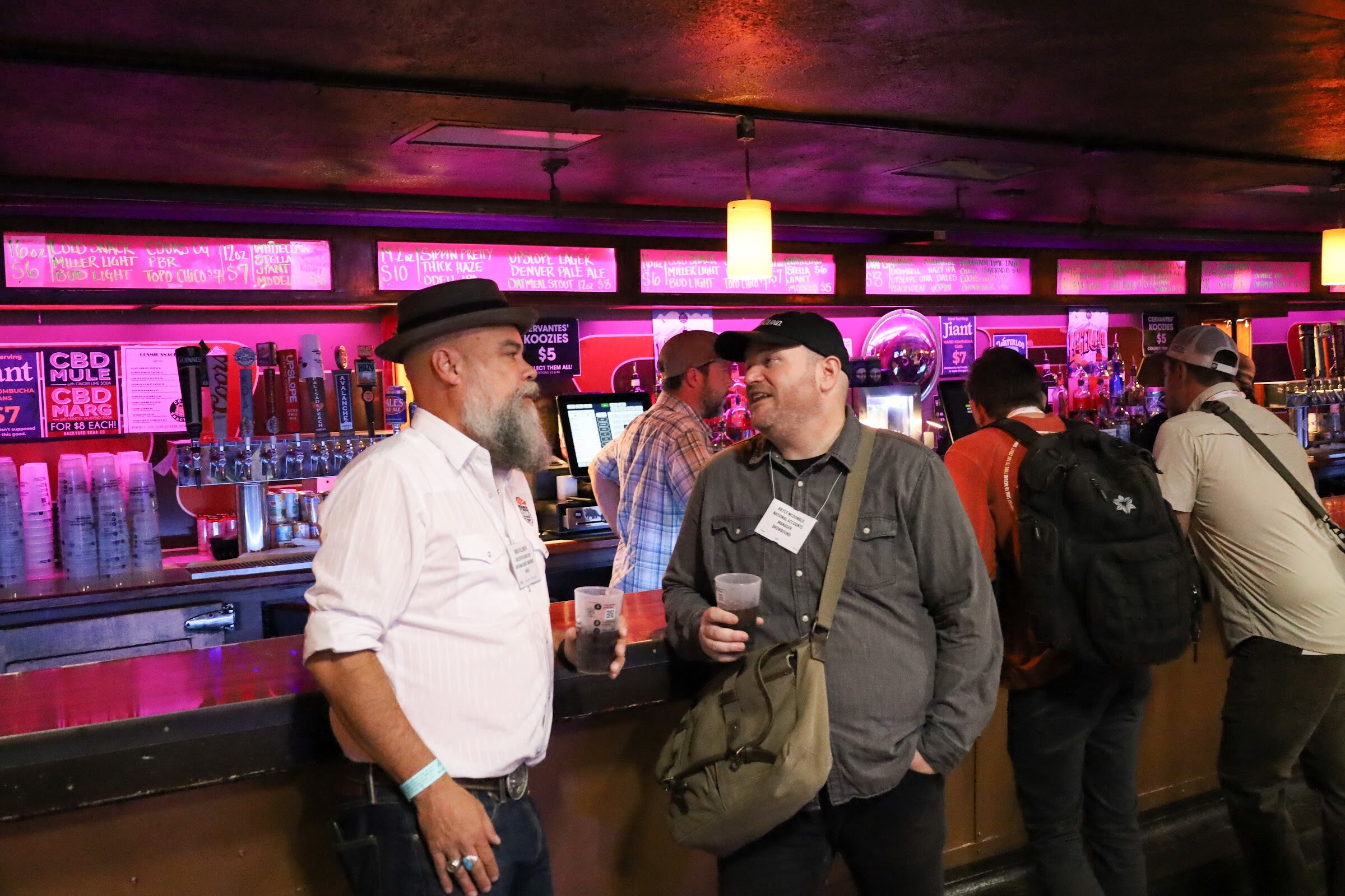 A man with a gray beard in a black hat and white shirt and a man in a black cap and gray button-up shirt with a canvas bag, both with name badges and plastic cups, talk at a bar with tap handles and chalkboard menus behind them.