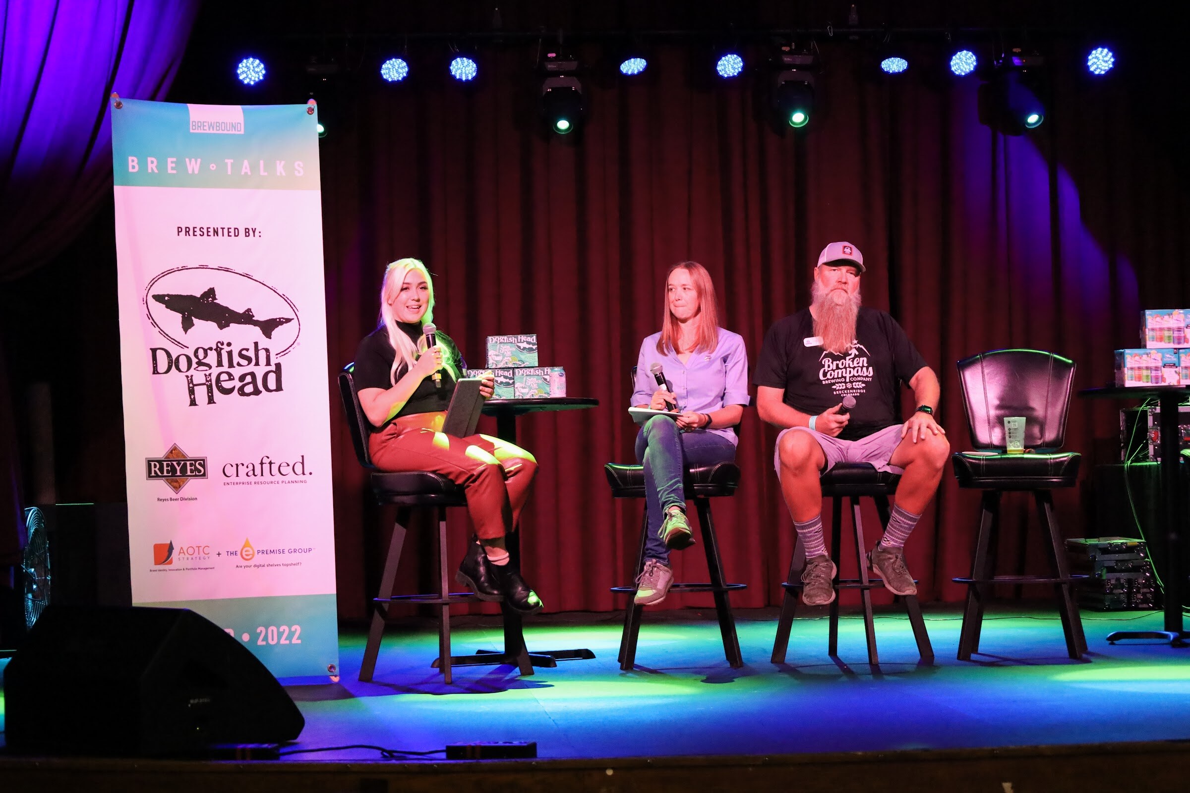 A woman with white-blonde hair in a black top and rust leather pants holds a microphone and a tablet, a woman in a lavender shirt holds a notepad, and a bearded man in a Broken Compass Brewing t-shirt sits on a bar stool, beside a Brew Talks banner.