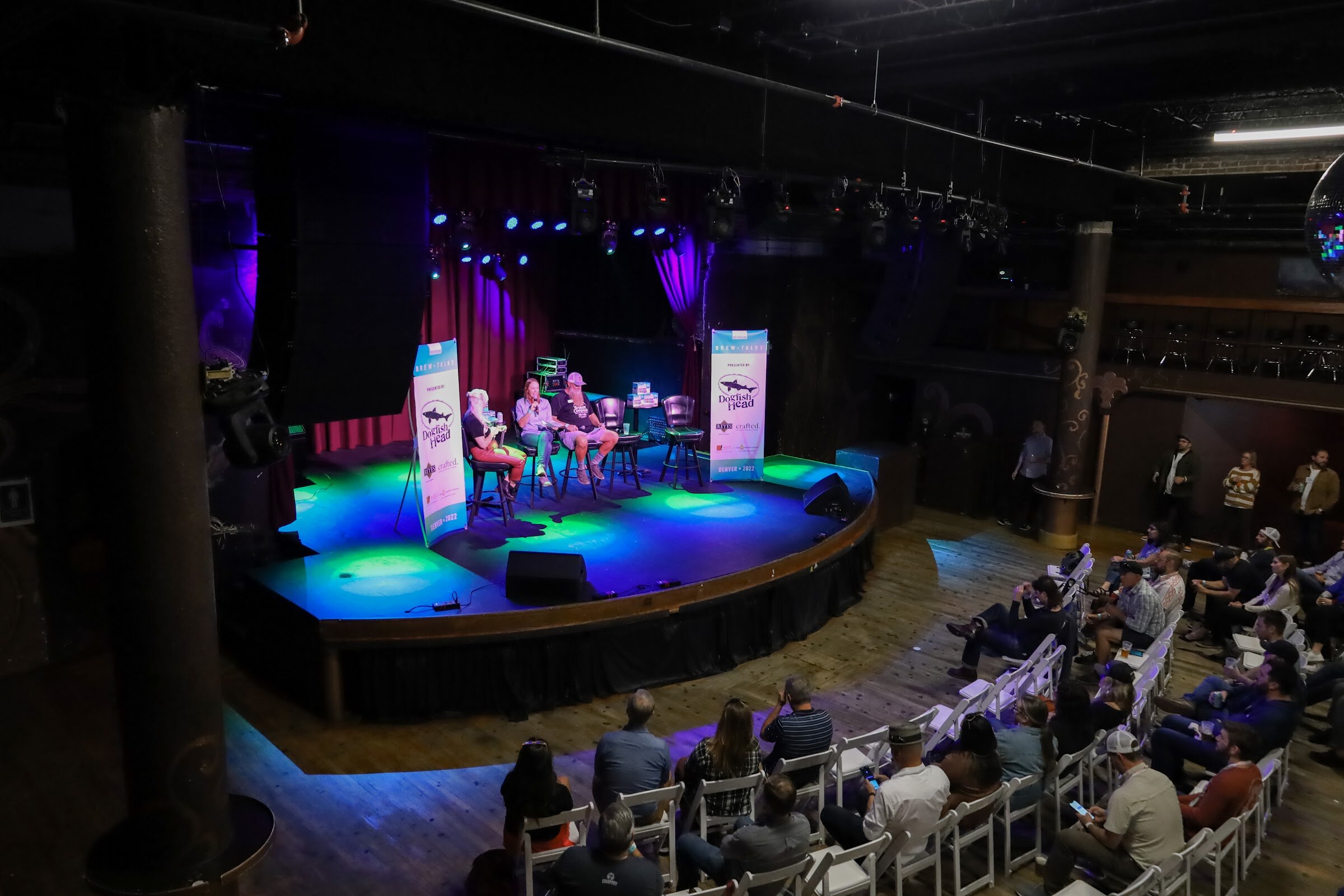 View from above of a curved stage lit in blue and green with three people seated on bar stools between two Brew Talks banners, with audience members in white folding chairs at the right.