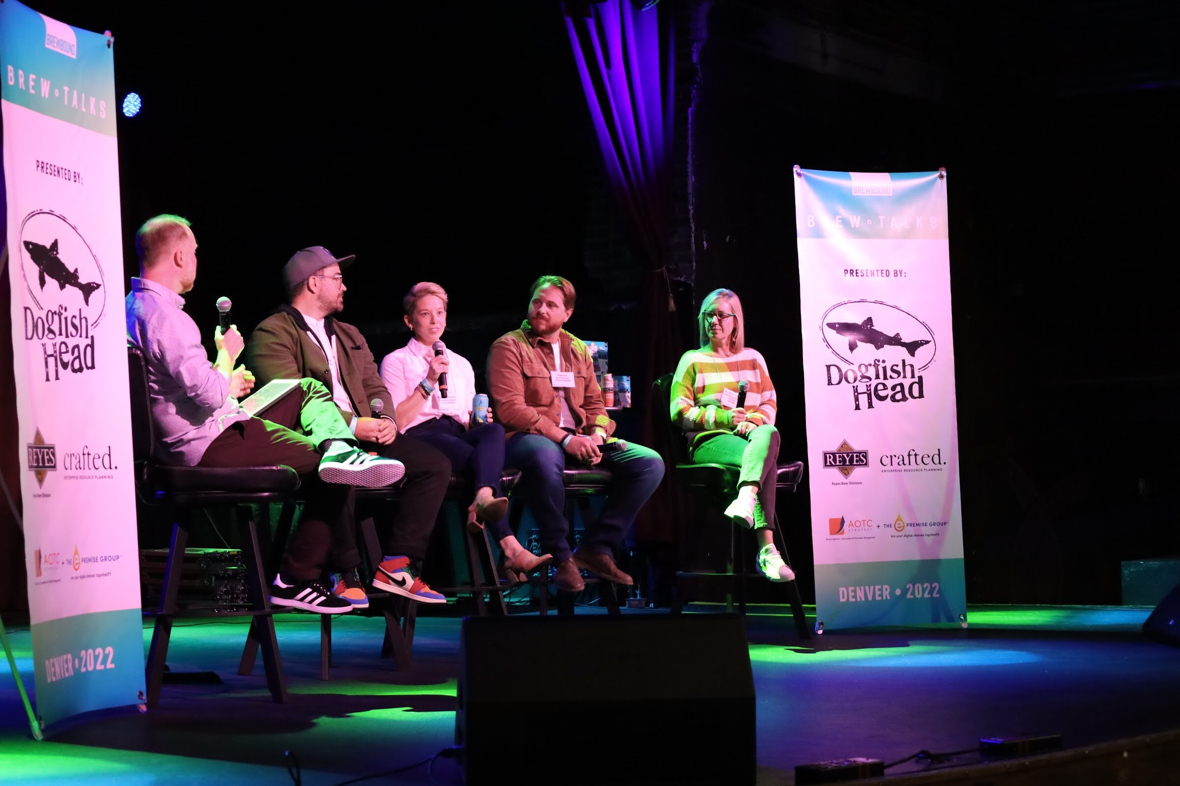 A man in a lilac shirt holds a microphone and a tablet and faces four panelists on bar stools, a man in a cap, glasses and olive jacket, a woman in a white shirt, a bearded man in a rust jacket and a woman in an orange-striped sweater, between two Brew Talks banners.