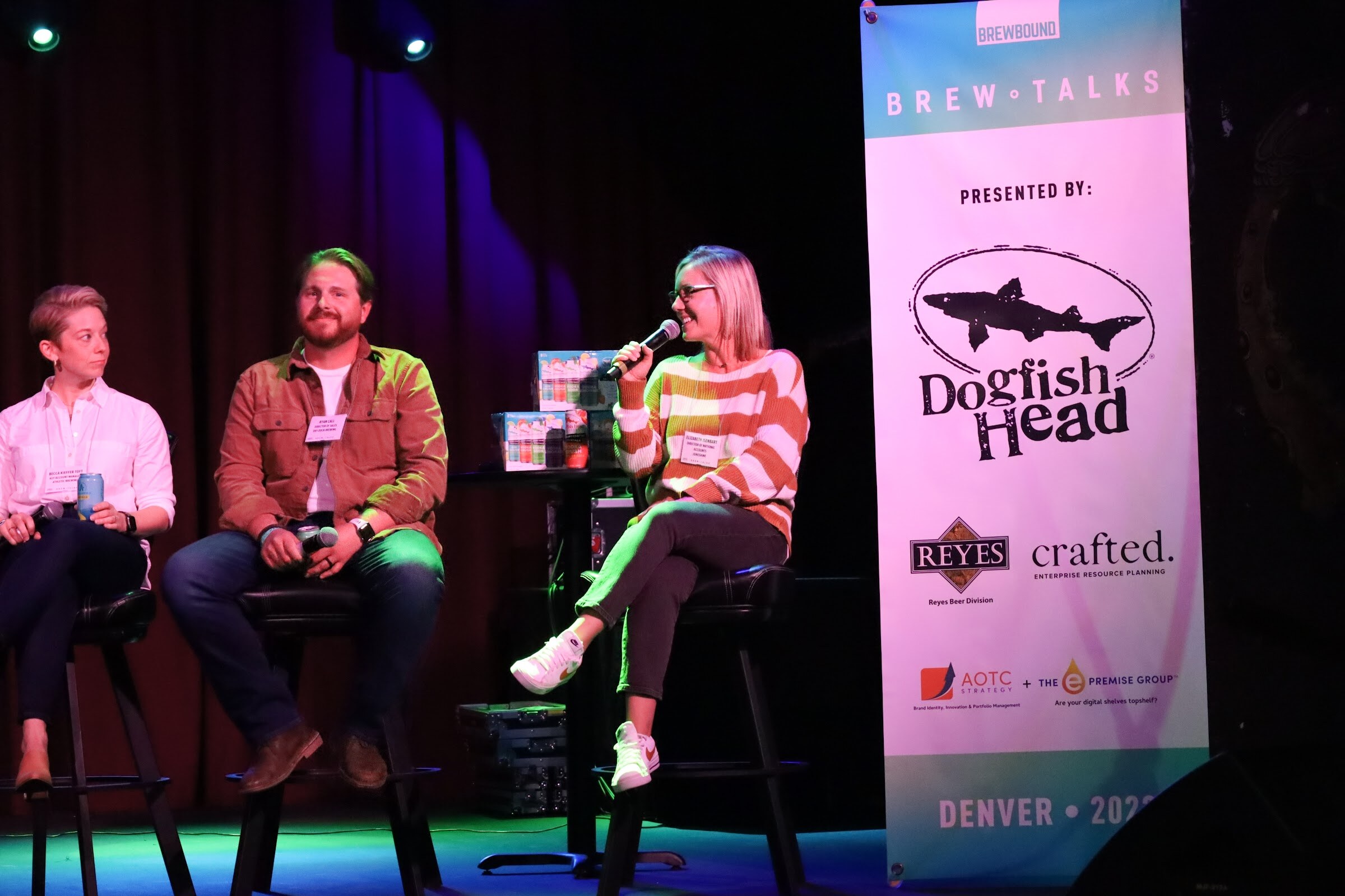 A blonde woman with glasses in an orange, white and green striped sweater speaks into a microphone while a bearded man in a rust jacket smiles and a woman in a white shirt looks on, beside a Brew Talks banner presented by Dogfish Head, Reyes and crafted.