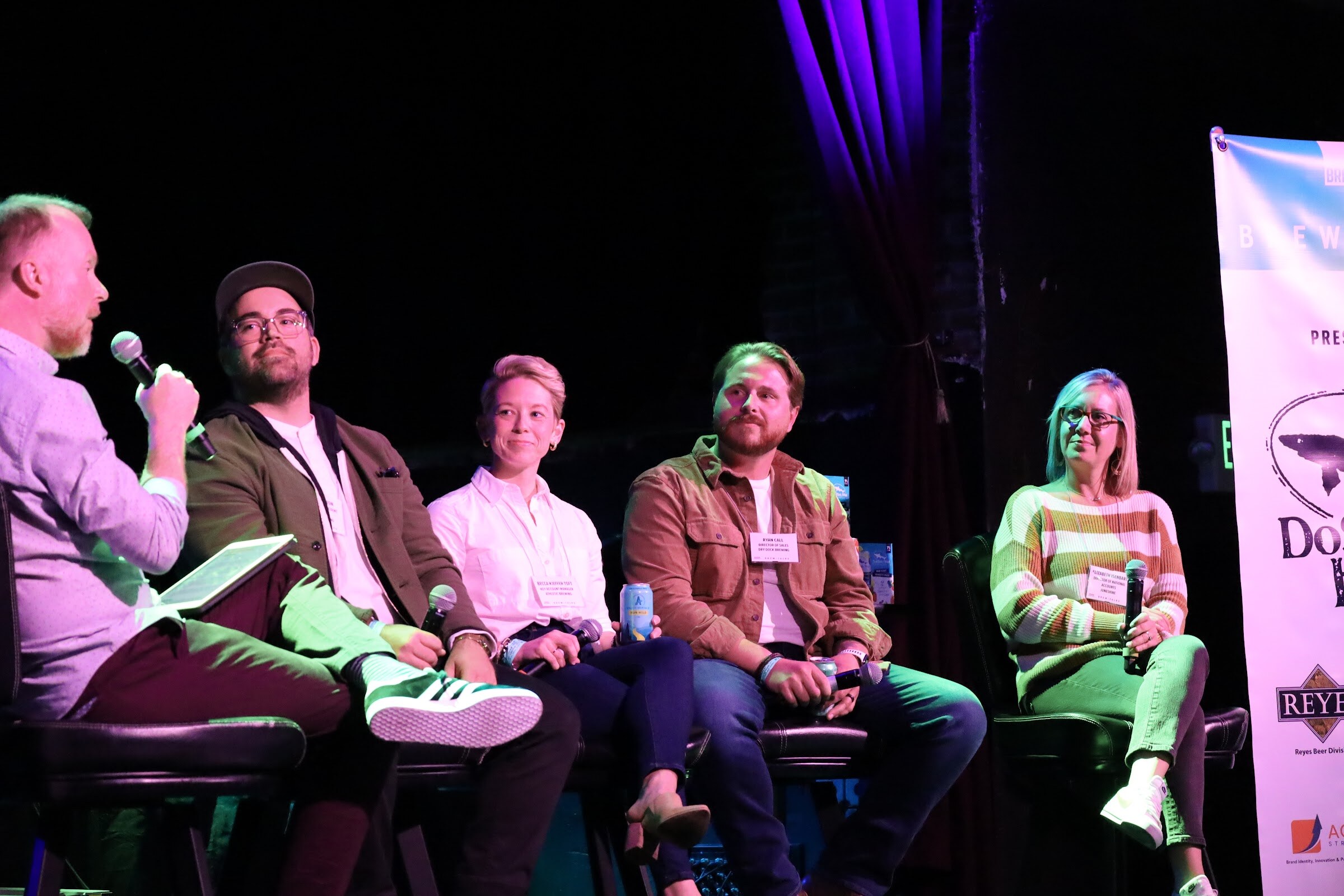 A man in a lilac shirt holding a tablet and microphone faces four panelists on bar stools, a man in a cap, glasses and olive jacket, a smiling woman in a white shirt holding a can, a bearded man in a rust jacket and a woman in a striped sweater.