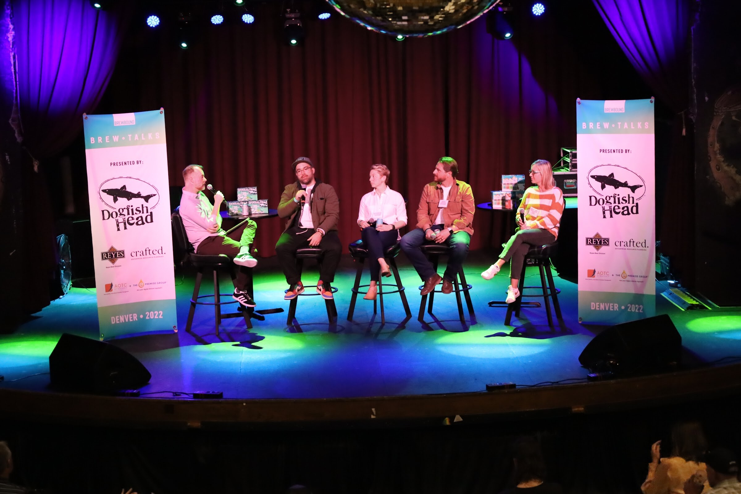 Five people on black bar stools on a stage lit in blue and green, with a disco ball at the top of the frame and a Brew Talks banner presented by Dogfish Head at each side.