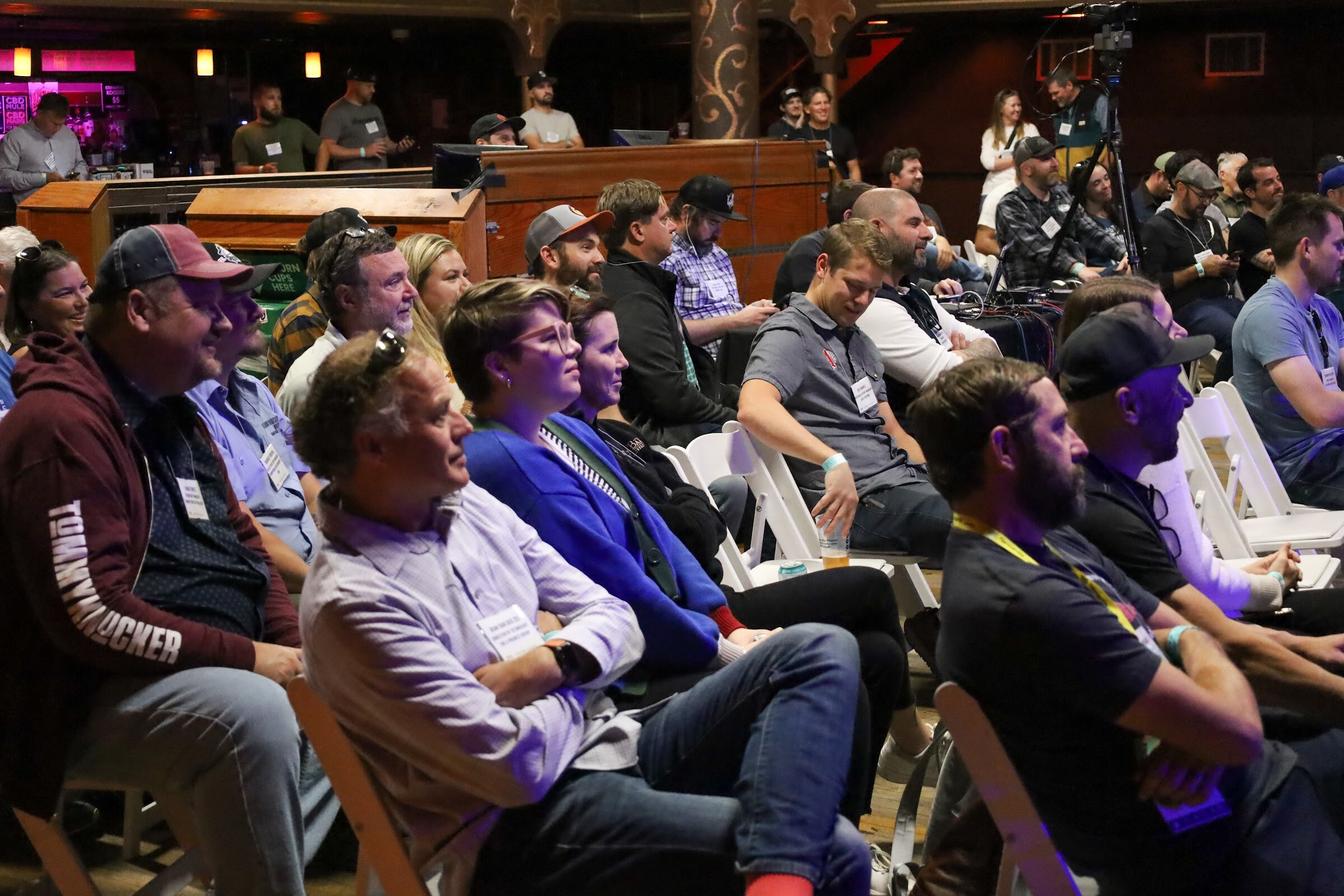 Close view of seated attendees with name badges listening, including a man in a maroon Tommyknocker hooded sweatshirt and cap, a woman in a blue cardigan and glasses and a man in a gray t-shirt, with the bar and standing guests behind them.