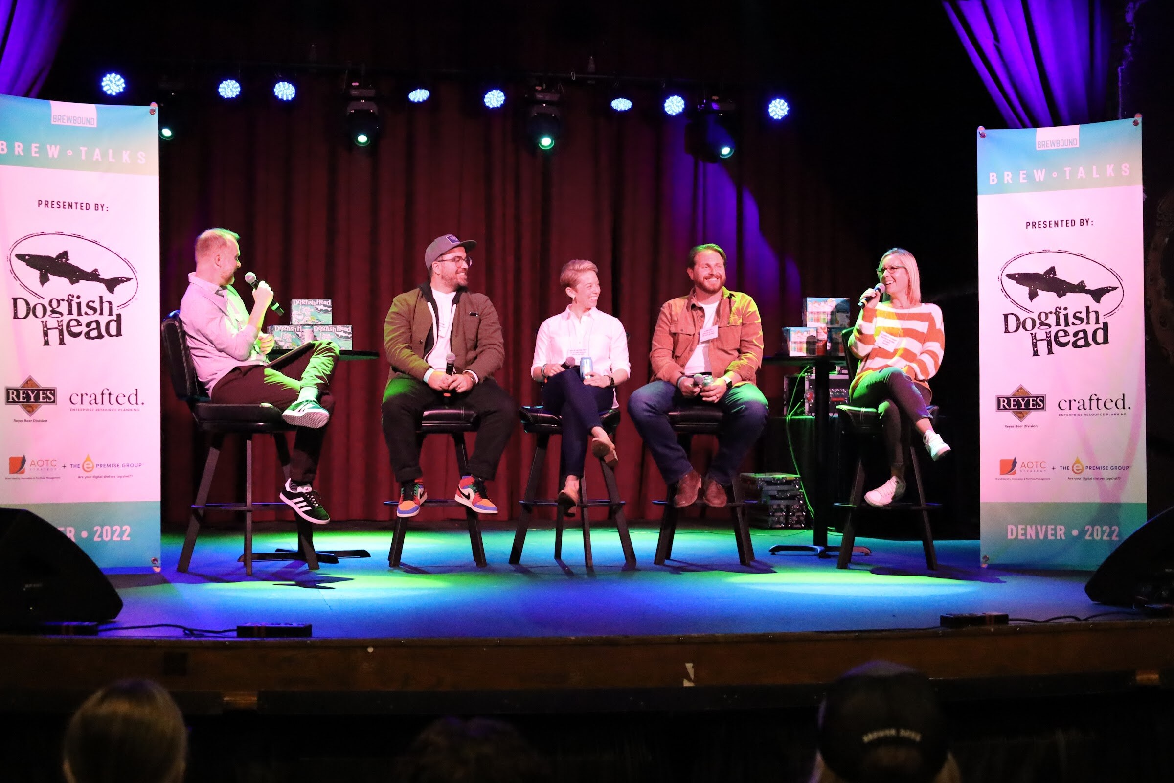 A man in a lilac shirt with a microphone and four panelists on black bar stools, including a woman in an orange-striped sweater speaking, sit in front of a red curtain between two Brew Talks banners.