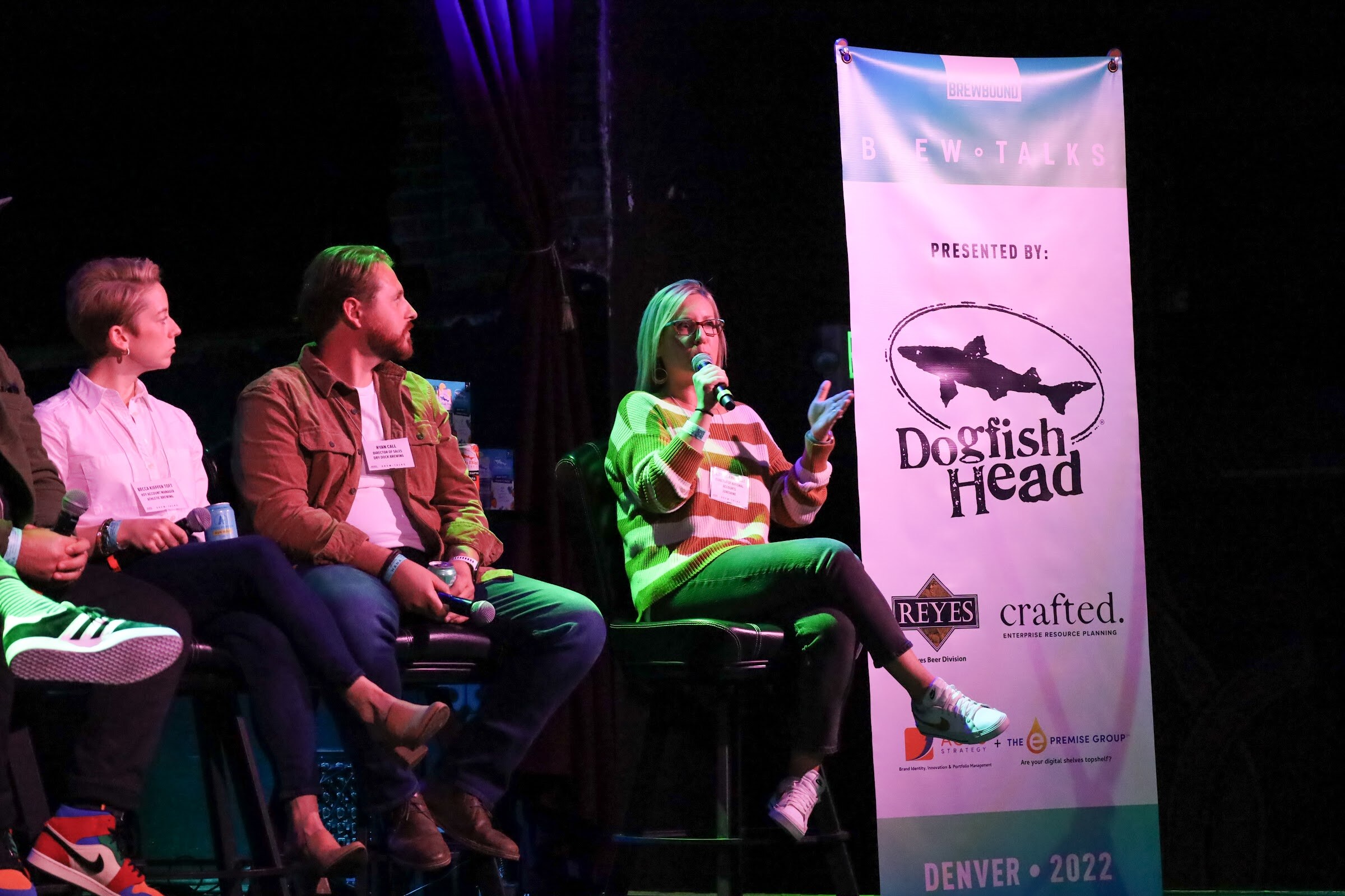 A woman with glasses in an orange, white and green striped sweater gestures with one hand as she speaks into a microphone, while a bearded man in a rust jacket and a woman in a white shirt sit beside her next to a Brew Talks banner.