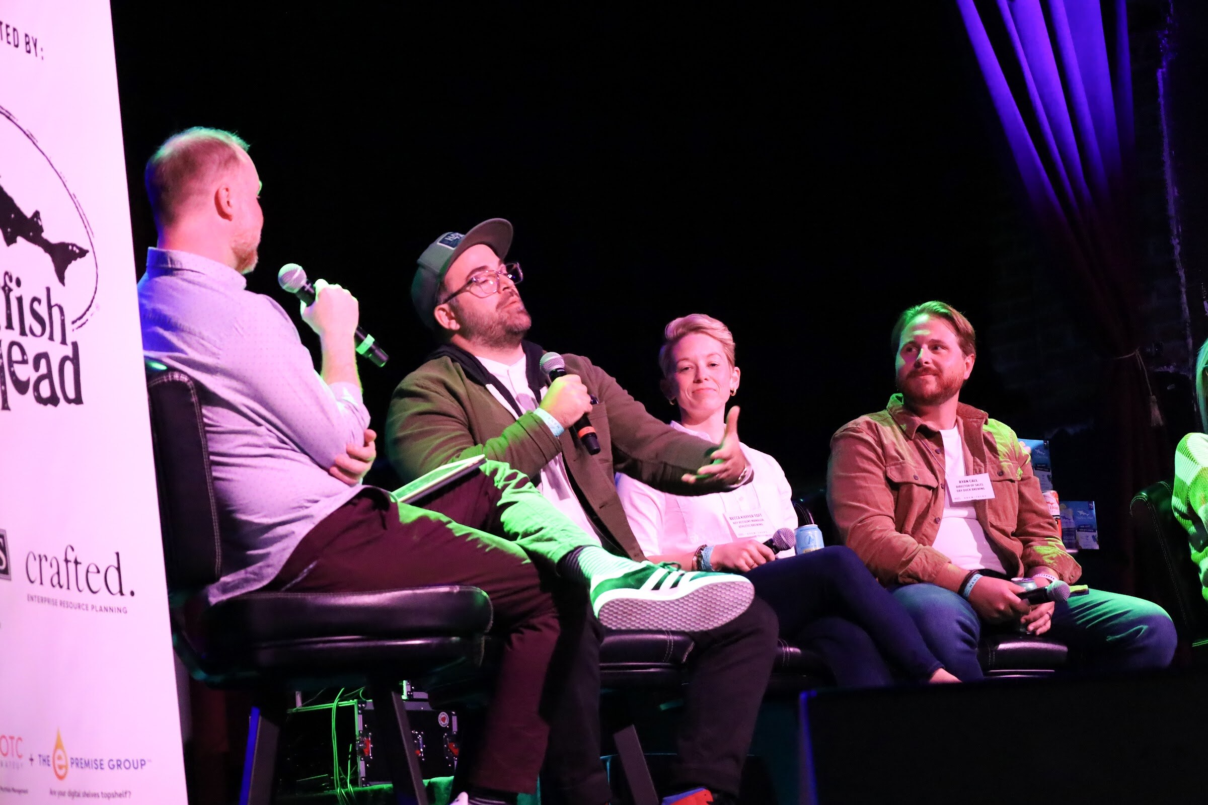 A man in a lilac shirt holds a microphone beside a panelist in a gray cap, glasses and olive jacket who gestures with one open hand, with a woman in a white shirt and a bearded man in a rust jacket seated next to him beside a Dogfish Head banner.