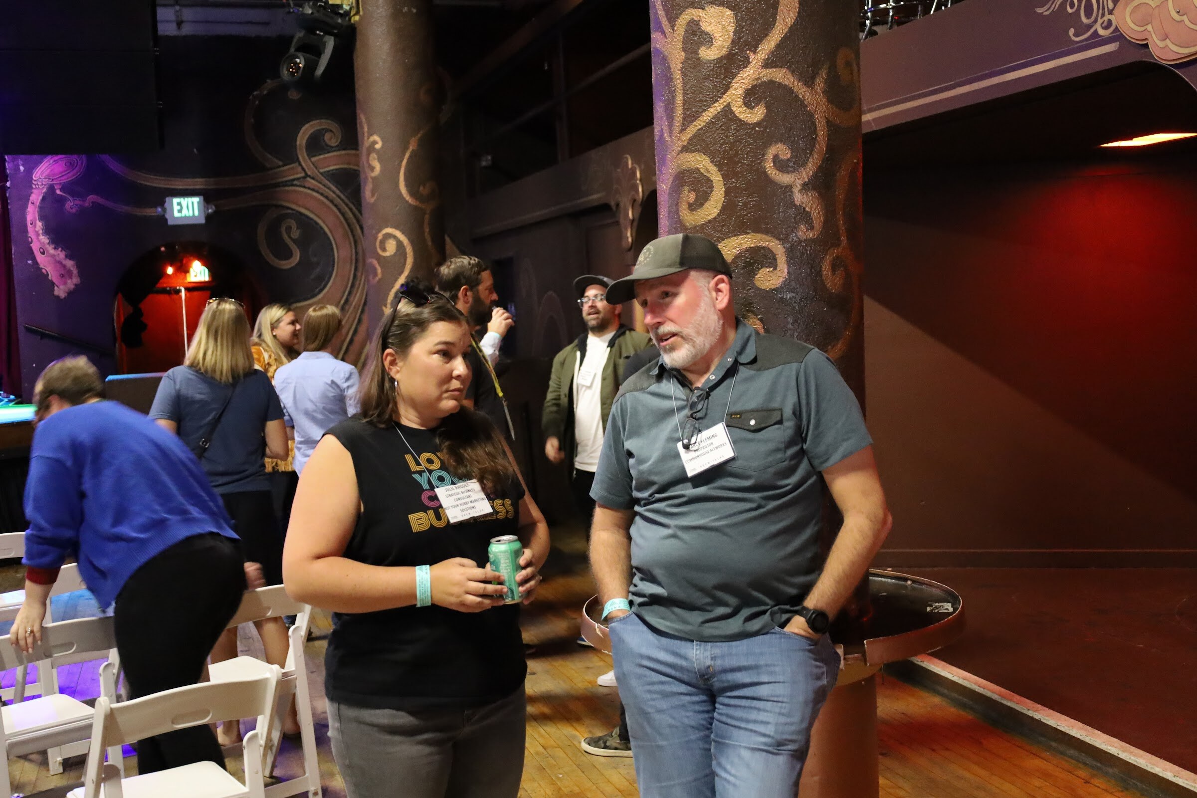 A woman in a black Love Your Craft Business tank top holding a green can and a man in a gray polo and dark cap, both wearing name badges, talk beside a painted pillar, with white folding chairs and other guests behind them.