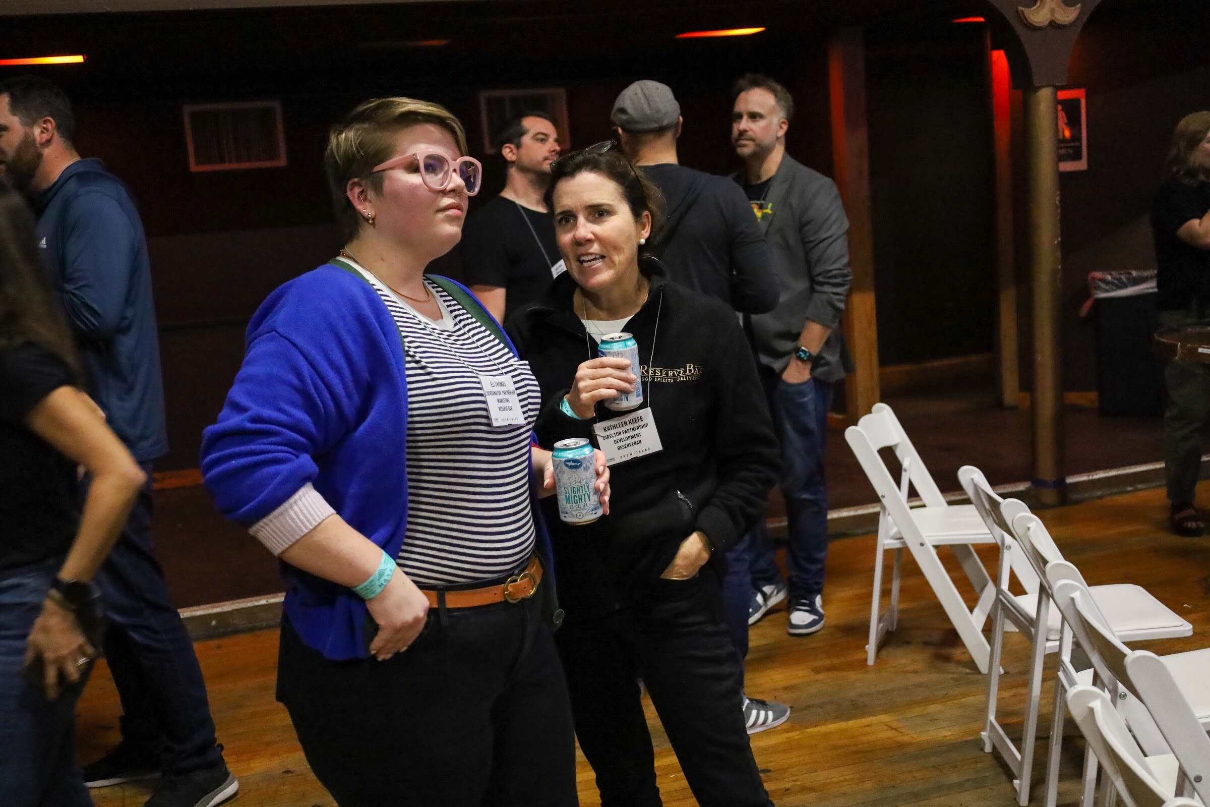 A person with pink glasses in a blue cardigan over a striped top and a woman in a black zip-up jacket each hold a can while talking, with white folding chairs and other guests behind them.
